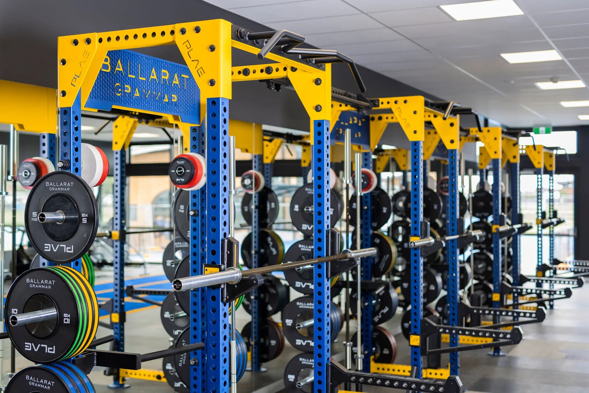 Row of blue-and-yellow PLAE ICON X power racks branded with the Ballarat Grammar name, loaded with PLAE bumper plates and barbells