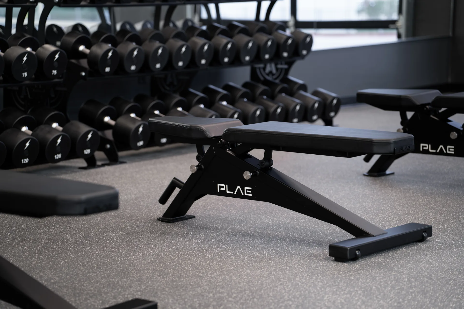 PLAE adjustable bench on grey rubber flooring beside a rack of PLAE dumbbells in the Bare Performance Nutrition gym