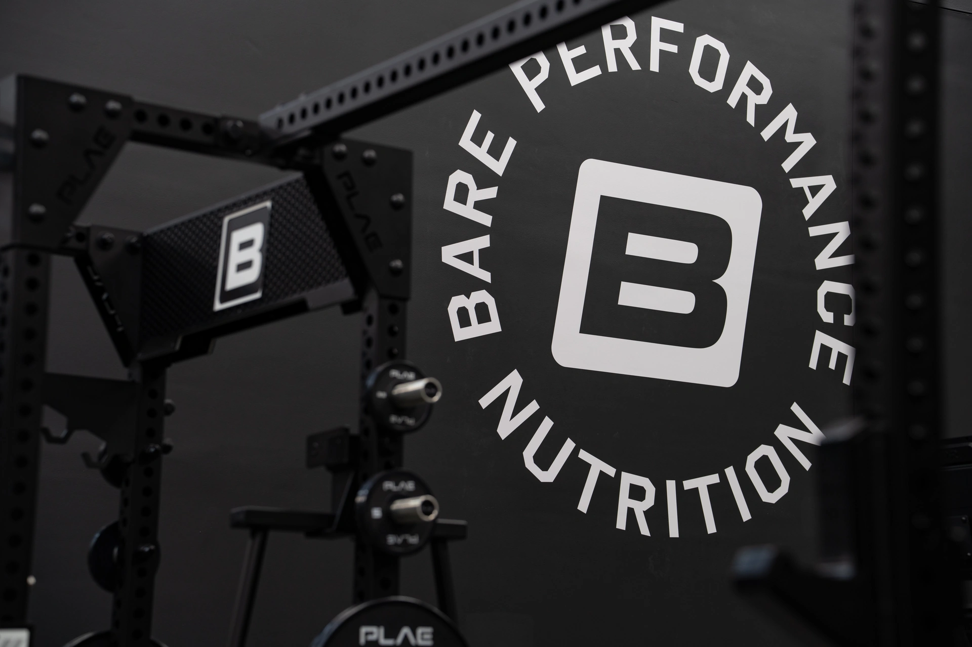 Bare Performance Nutrition circular logo on the wall beside a black PLAE ICON X rack and PLAE bumper plates