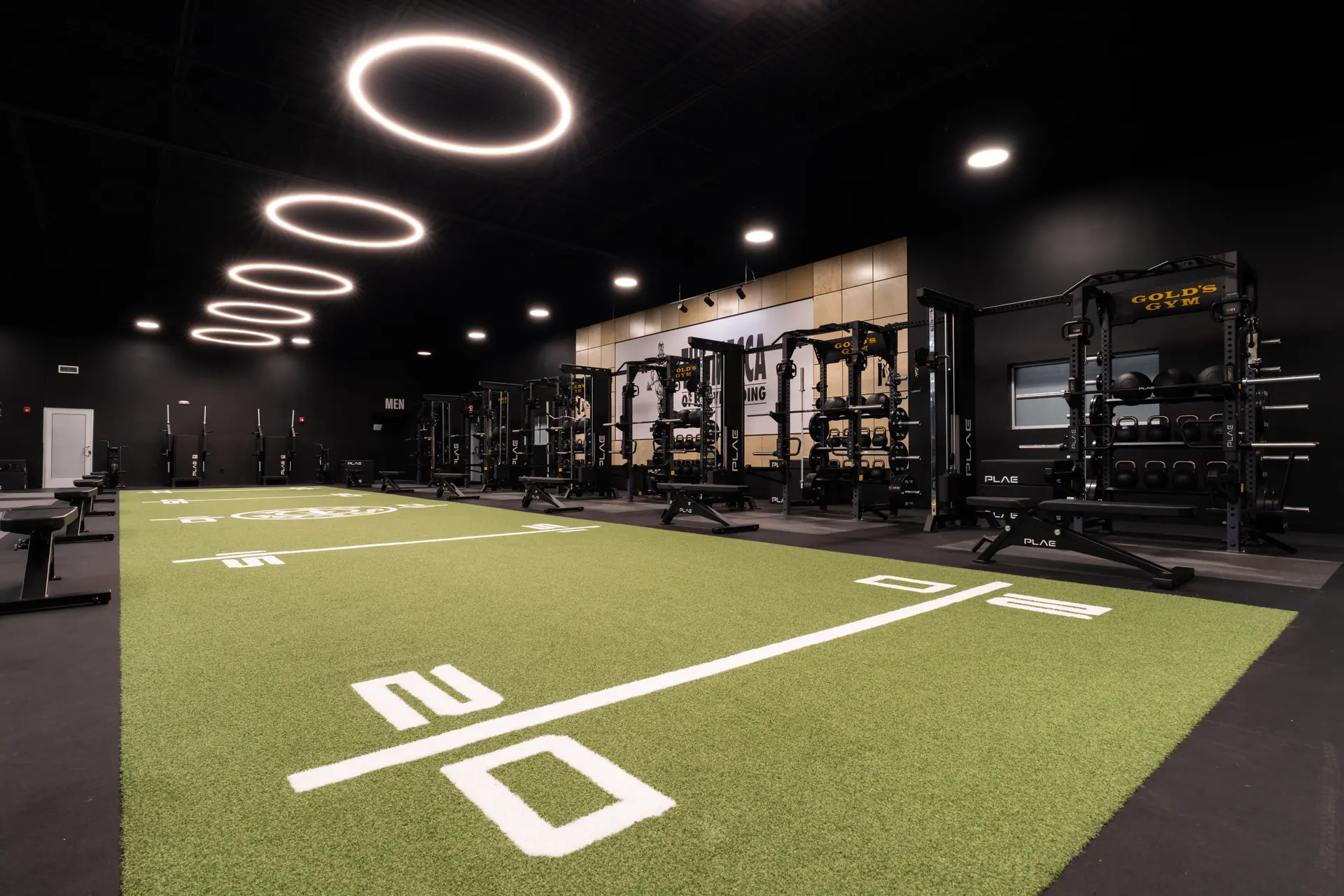 Row of Gold's Gym PLAE ICON X racks stocked with slam balls and kettlebells beside the green ATTACK turf track in Austin