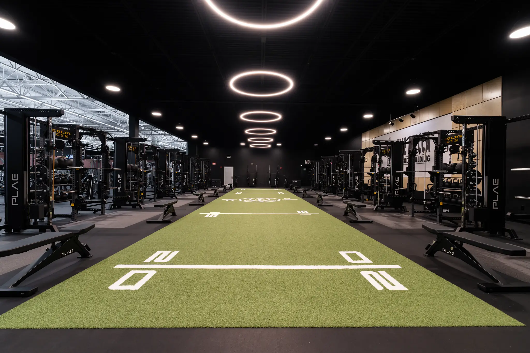 Gold's Gym Austin strength floor with PLAE ICON X racks and benches flanking the green ATTACK turf track beneath halo ring lights