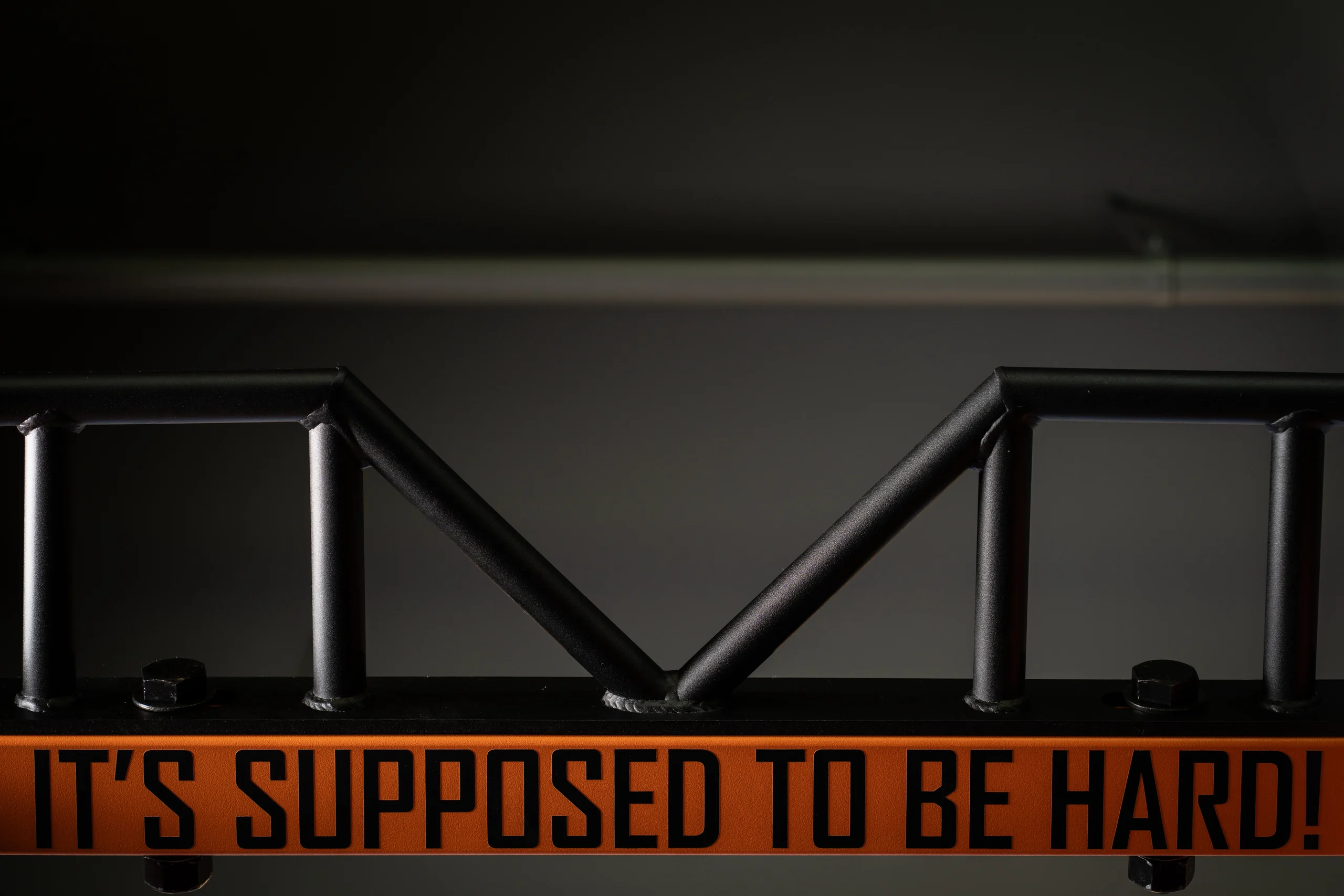 PLAE rack crossmember with black-on-orange lettering reading "It's Supposed To Be Hard" above a welded steel pull-up truss in the Tennessee golf facility.
