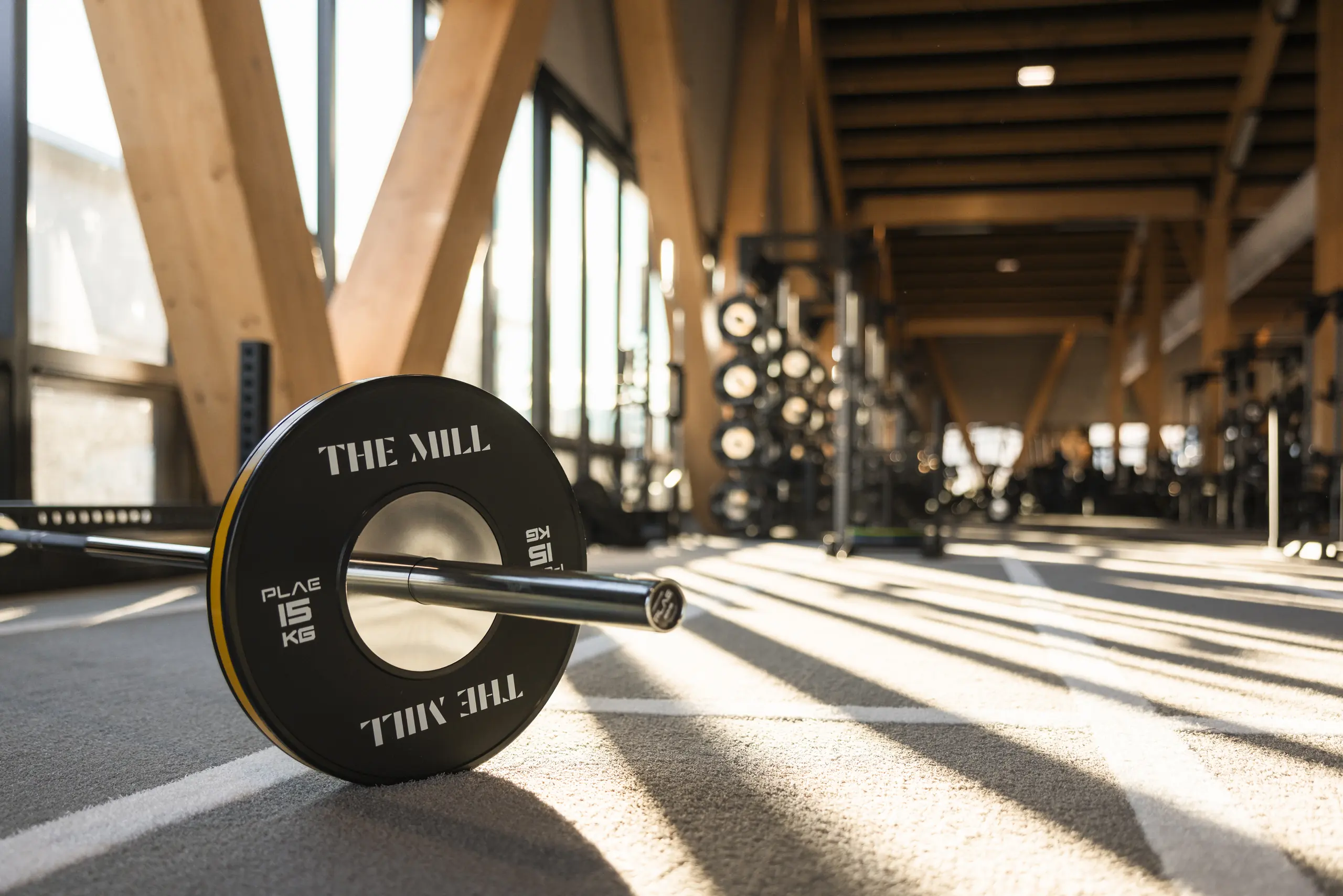 Custom "The Mill" 15kg PLAE bumper plate and barbell in low sun on Achieve rubber flooring, with ICONx racks and timber columns behind at The Mill, Queenstown.