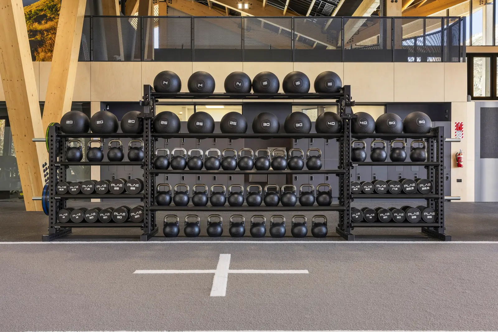 PLAE ICONx storage rack holding slam balls, kettlebells and round dumbbells beside a cross marking inlaid into Achieve rubber flooring at The Mill.
