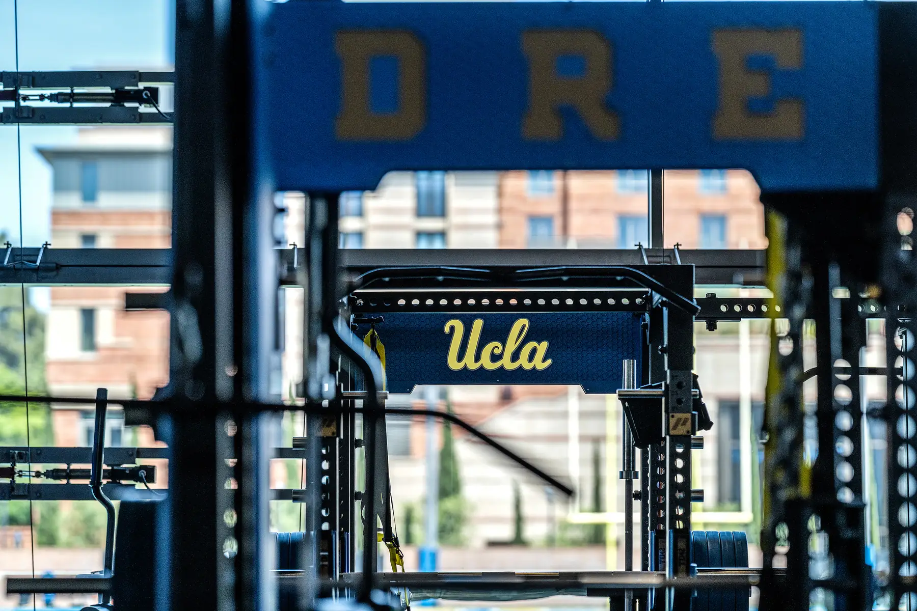 View through gym equipment framing a UCLA-branded PLAE power rack with the training field beyond.