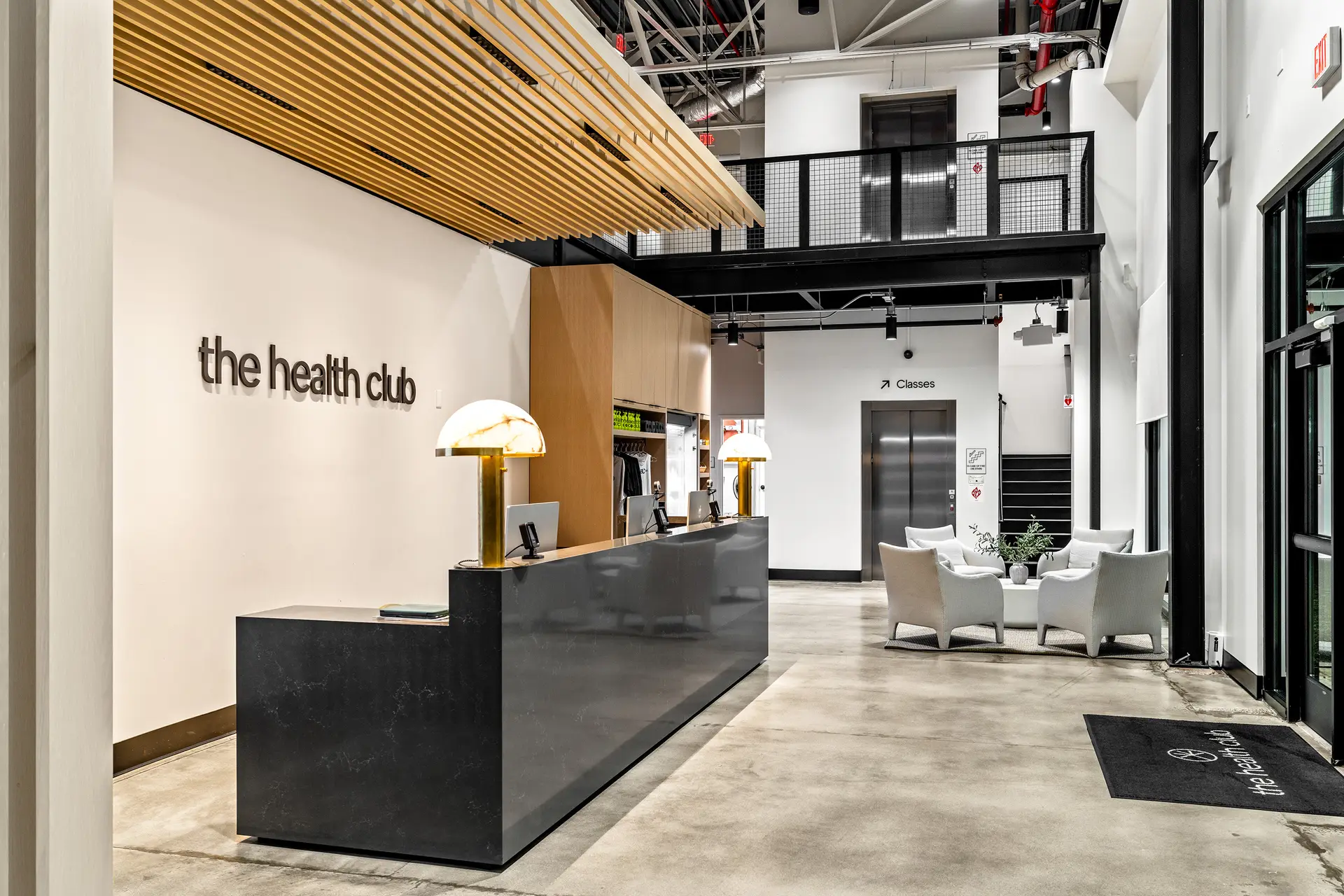 The Health Club reception desk with backlit signage, alabaster lamps and a lounge seating area beside the elevator and Classes stairway.