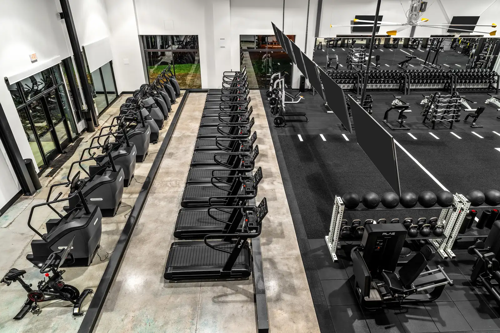 Overhead view of a row of PLAE treadmills beside Precor stair climbers and a Peloton bike, with a PLAE leg press and dumbbell racks on the turf floor.