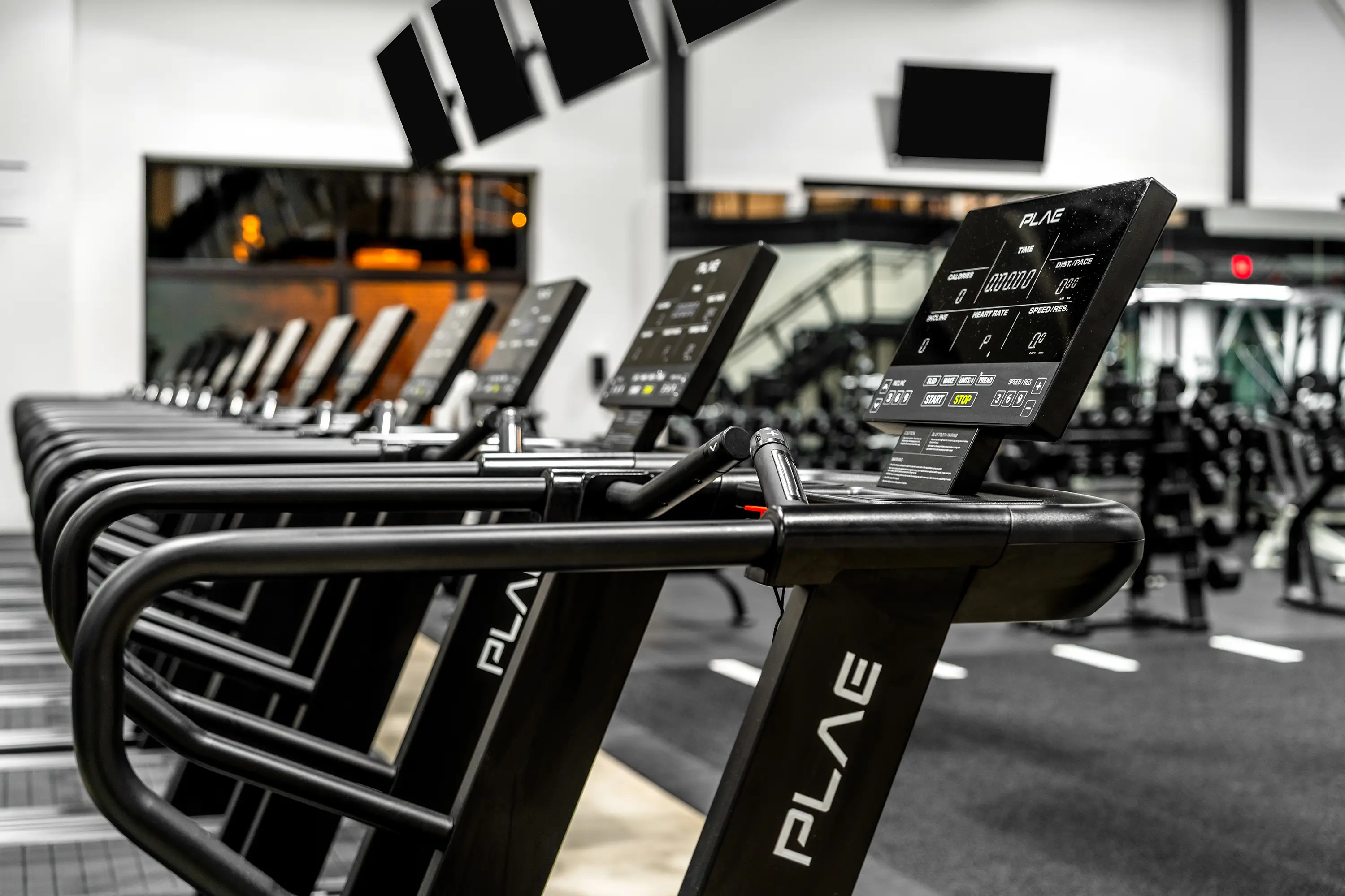 Detail of PLAE treadmill consoles and branded handrails in a lineup of PLAE cardio machines, with dumbbell racks softly blurred behind.