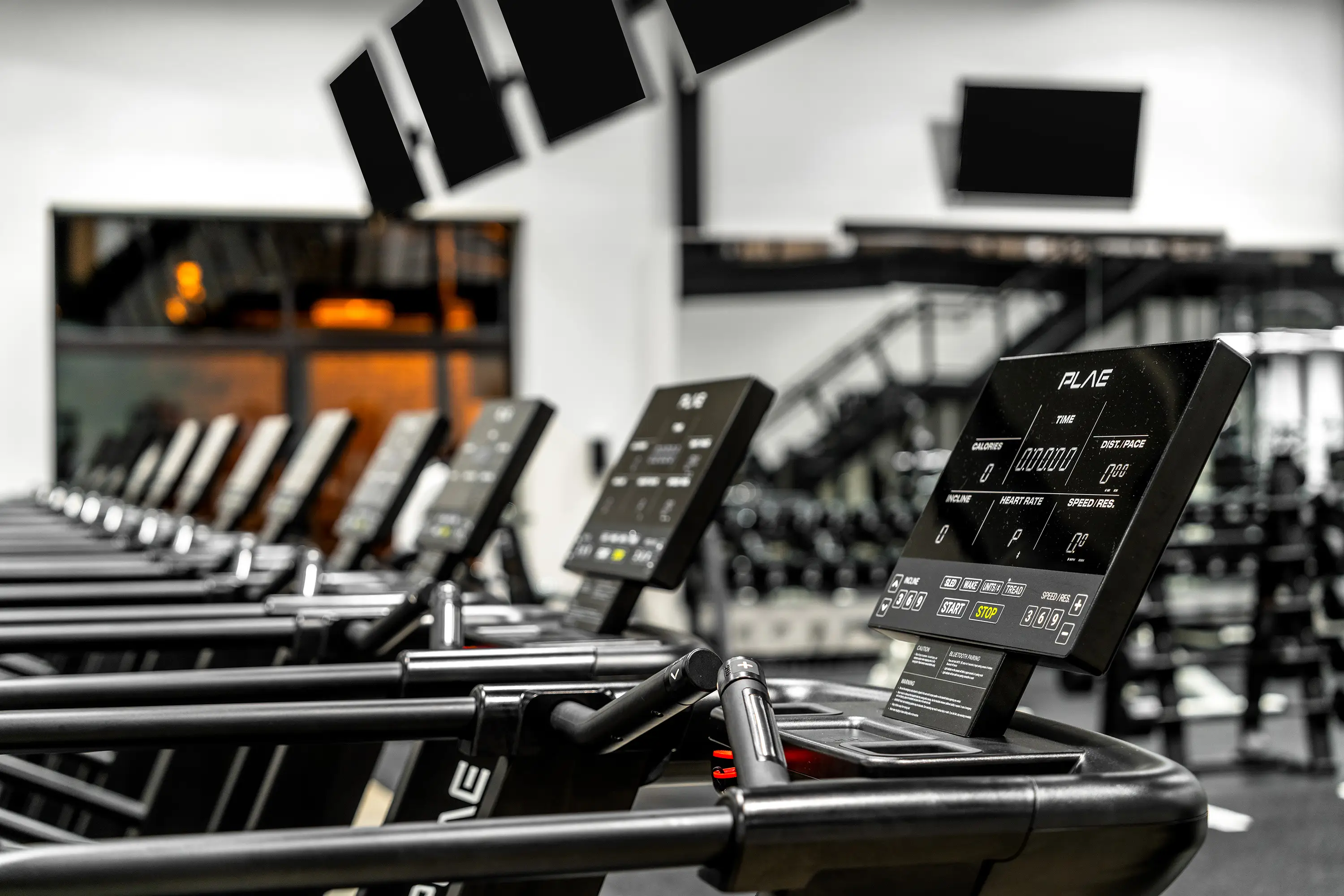 Close-up of a PLAE treadmill console showing the time, calories and distance display, with a full row of PLAE treadmills receding behind.