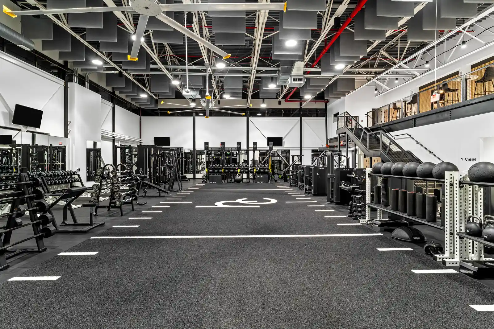 Turf strength floor looking toward PLAE functional training machines and rigs, flanked by slam balls, kettlebells and dumbbells beneath the Classes mezzanine.
