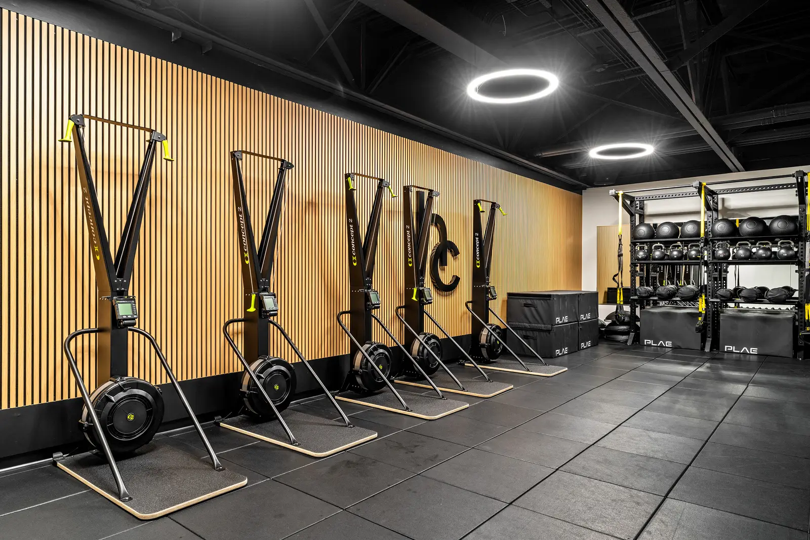 Row of Concept2 SkiErgs mounted along the wood-slat wall next to branded PLAE plyo boxes and a kettlebell and slam ball rack.
