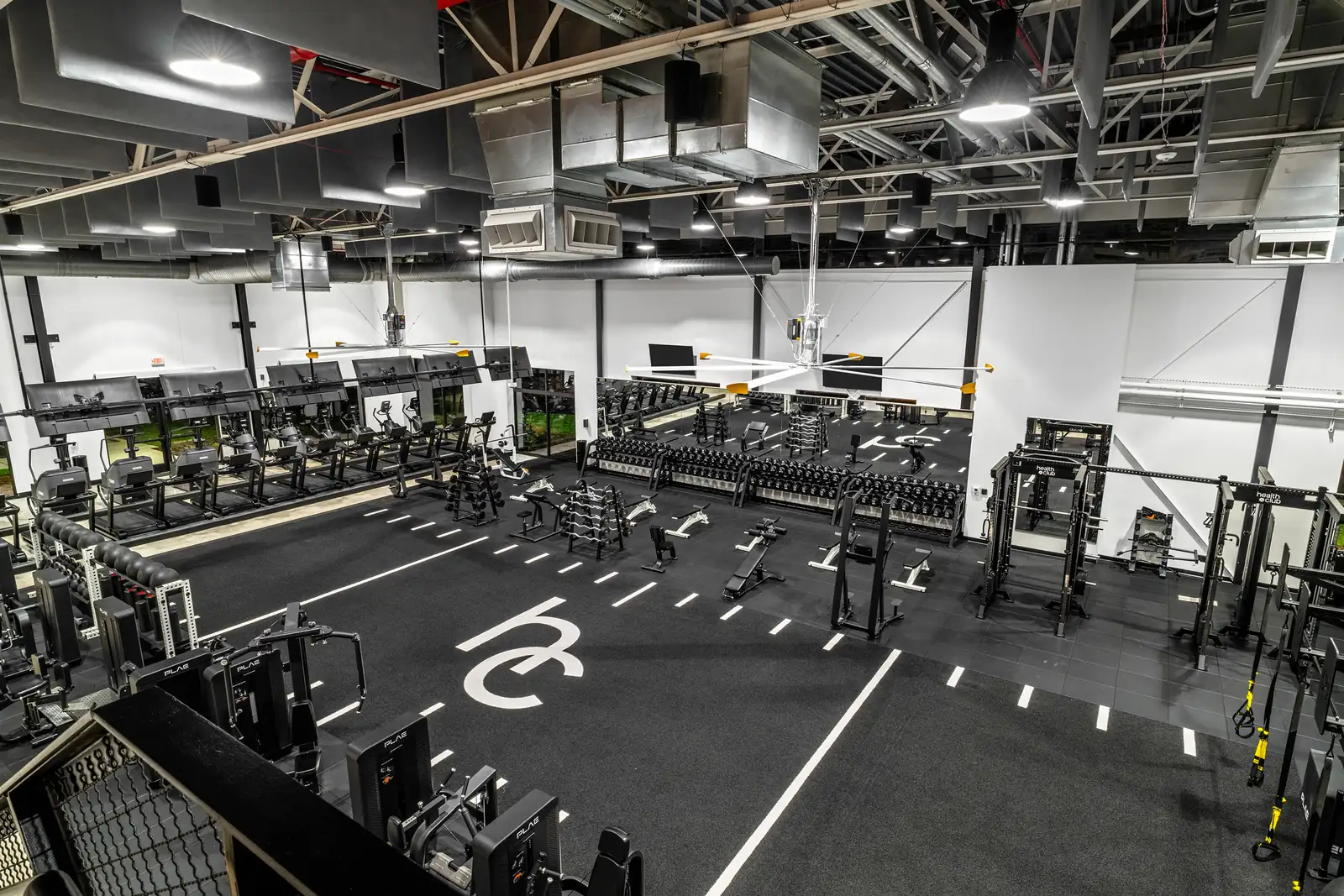 Aerial view of the free-weight zone with PLAE selectorized machines, health club power racks, tiered dumbbell rows and treadmills lining the mirrored wall.