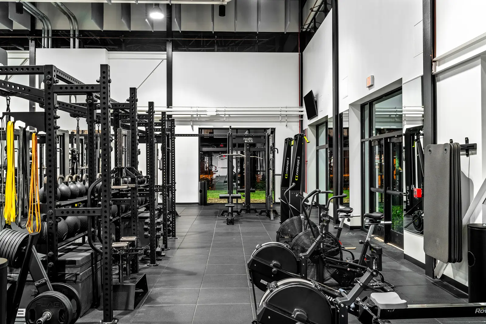 Rig-lined walkway with PLAE bumper plates and barbells, Concept2 SkiErgs, Assault bikes and a "health club" branded rack framing the far doorway.