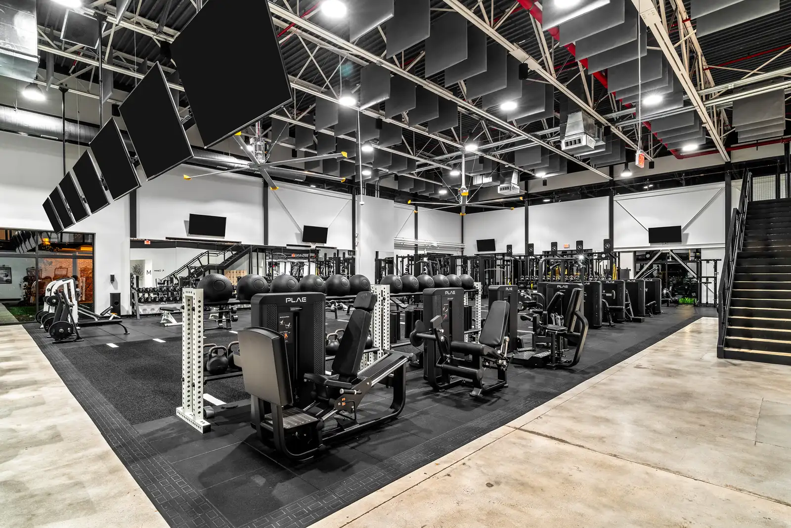 PLAE seated leg press and selectorized strength machines on the main training floor at The Health Club, with a slam ball rack, kettlebells and hanging acoustic panels overhead.