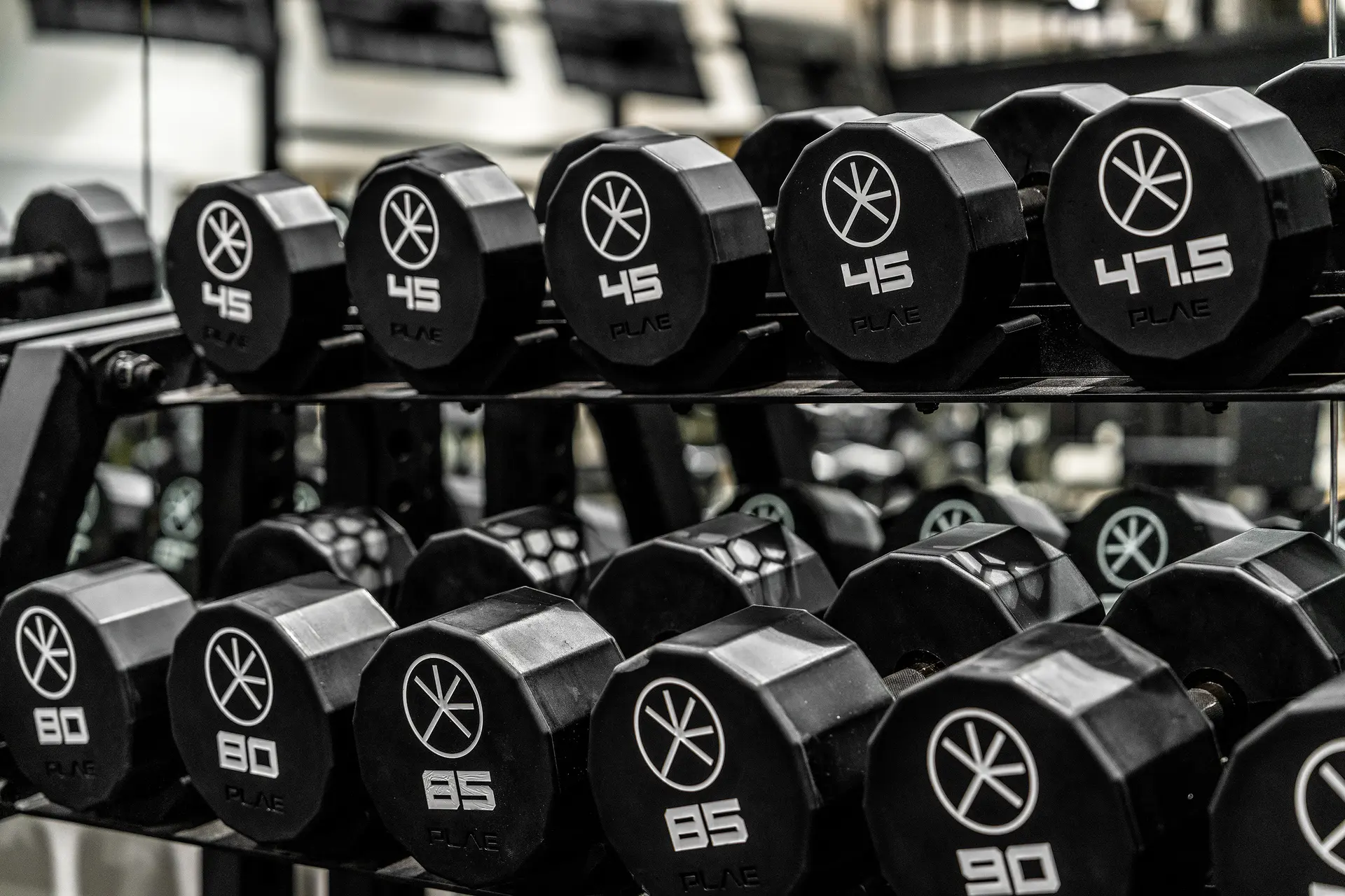 Close-up of PLAE urethane dumbbells on the rack — 45, 47.5, 80, 85 and 90 lb — showing the PLAE asterisk logo on each end cap.