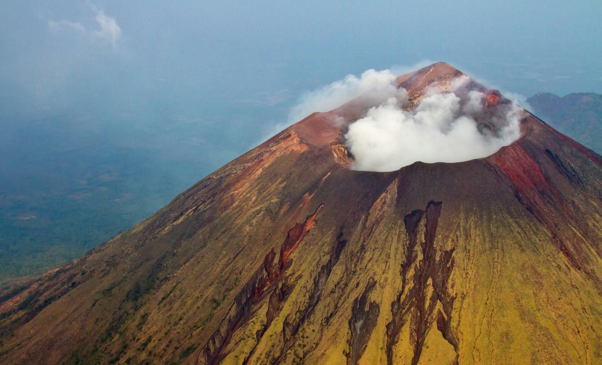 Steam rising from the crater of a volcanic cone with red and ochre slopes
