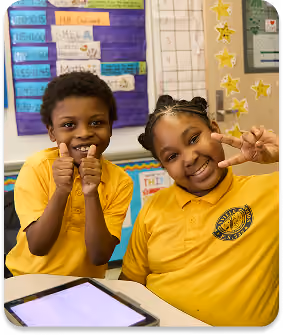 Two smiling students in yellow uniforms giving thumbs up in classroom