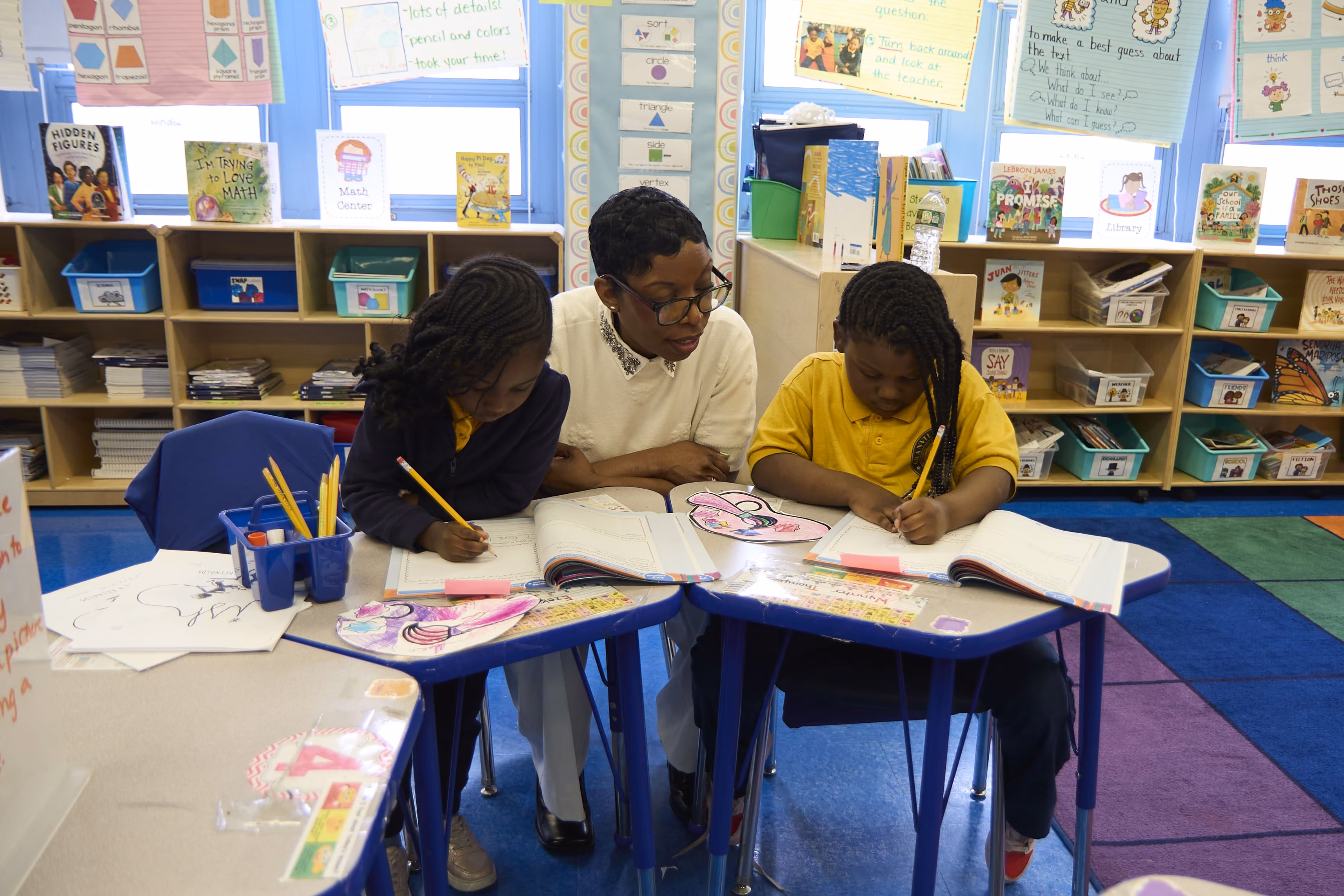 Teacher helping two students write in colorful elementary classroom