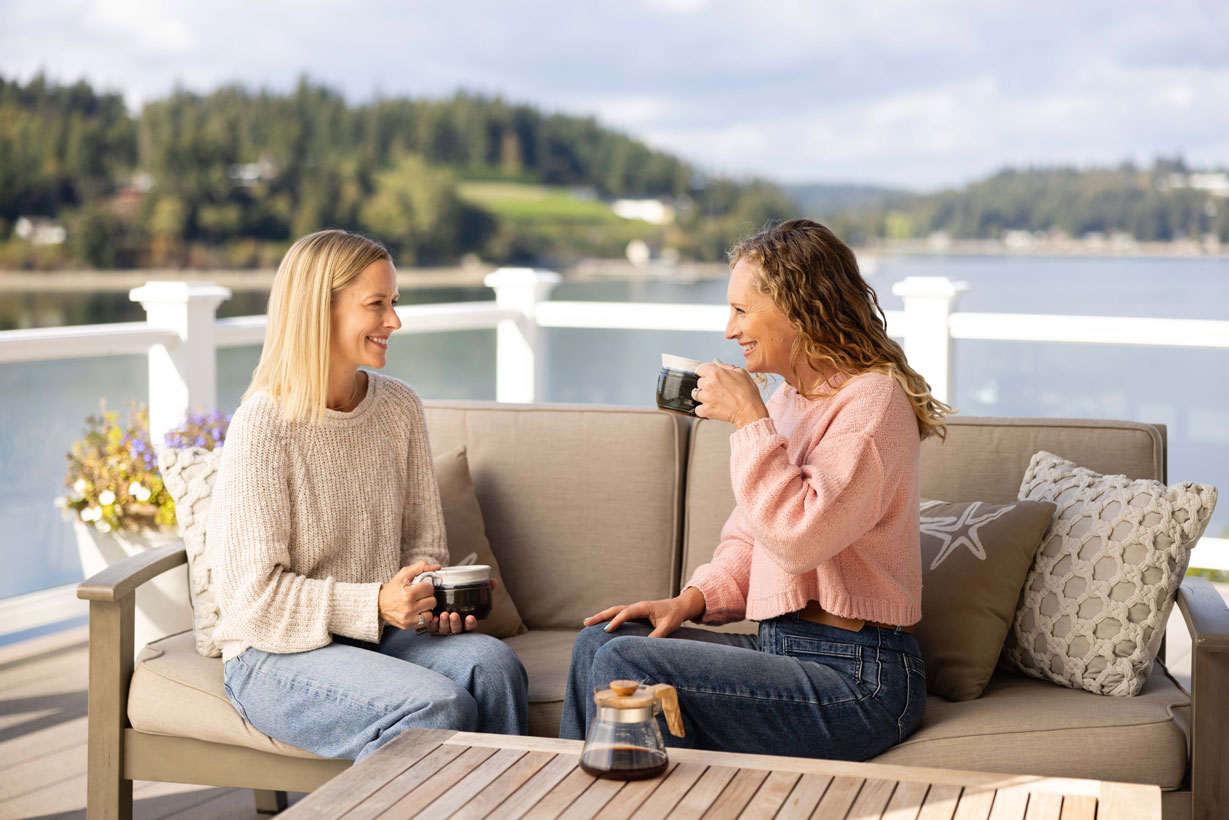 Two women enjoying locally roasted Cutters Point Coffee together on a deck overlooking the Gig Harbor waterfront, relaxing and talking on a sunny Pacific Northwest morning.