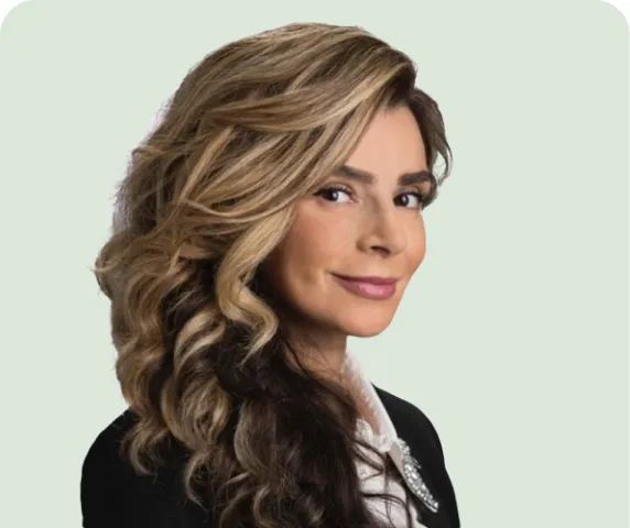 Smiling woman with long wavy highlighted hair wearing a black blazer and white blouse against a light green background.