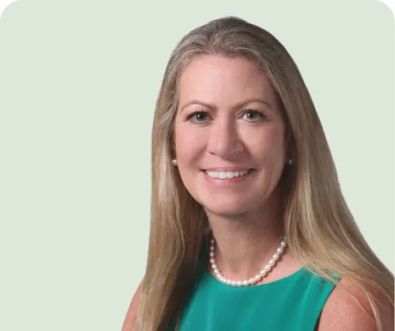 Smiling woman with long blonde hair wearing a green sleeveless top and a pearl necklace against a plain light background.
