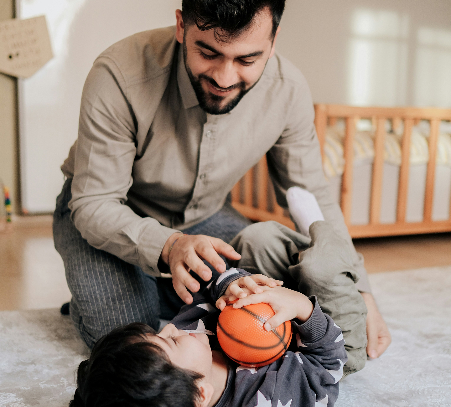 A father playing with his young child on a rug, both holding an orange basketball, with a crib visible in the background, promoting a joyful family moment.