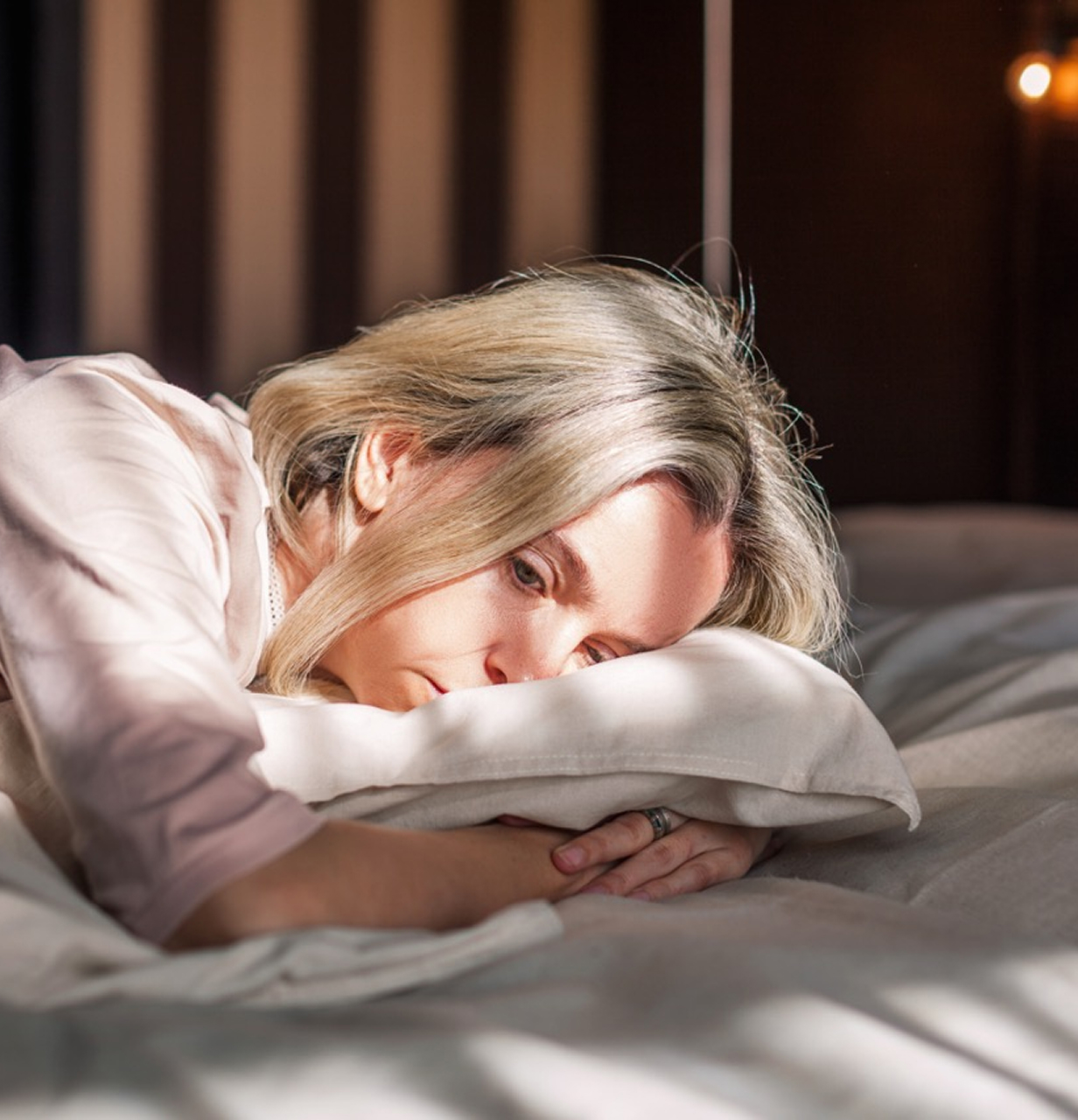 A woman lying in bed, resting her head on a pillow with a tired expression, illustrating the impact of headache or migraine symptoms.