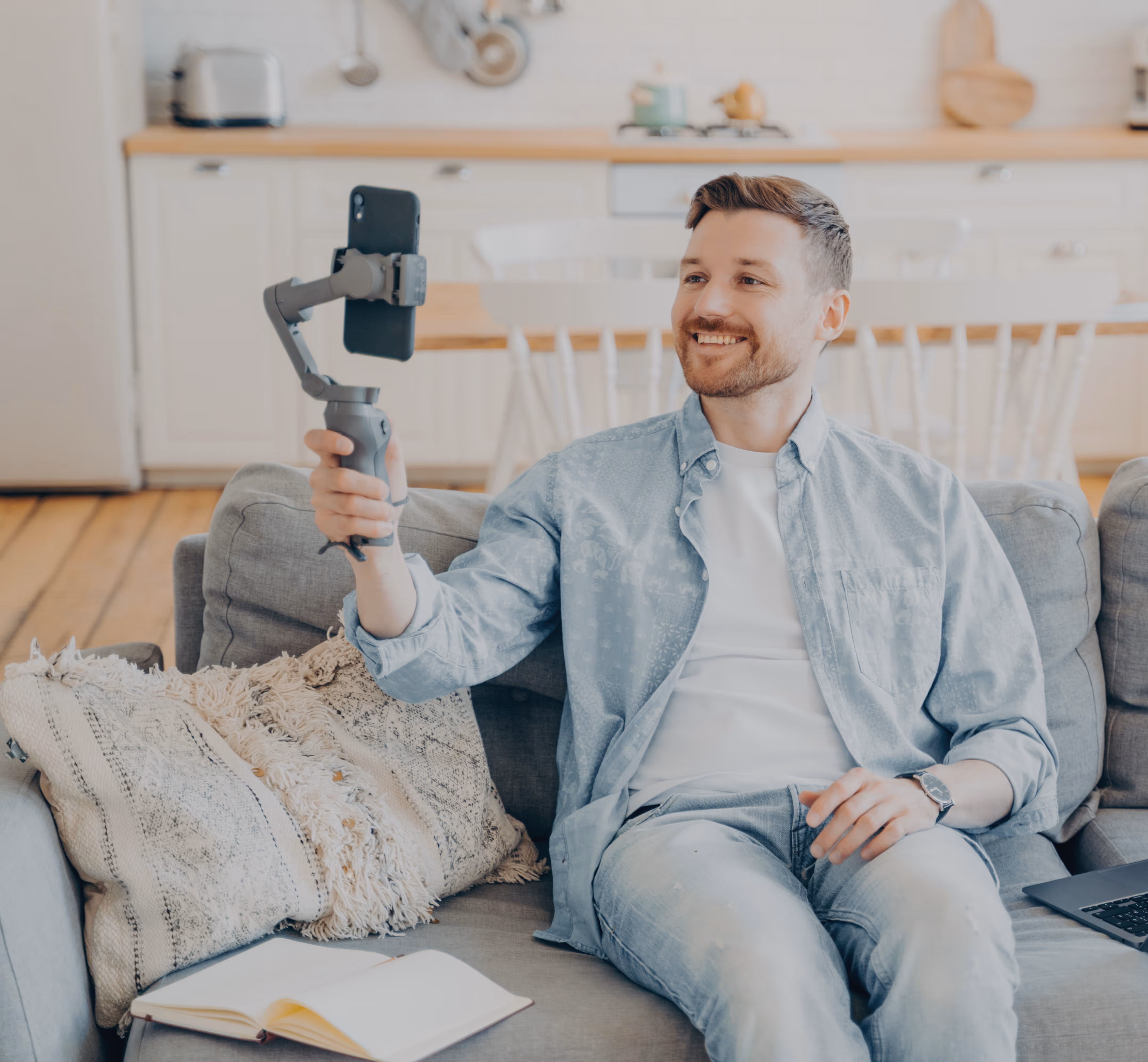 Smiling man sitting on a couch in a living room, holding a smartphone on a gimbal stabilizer.