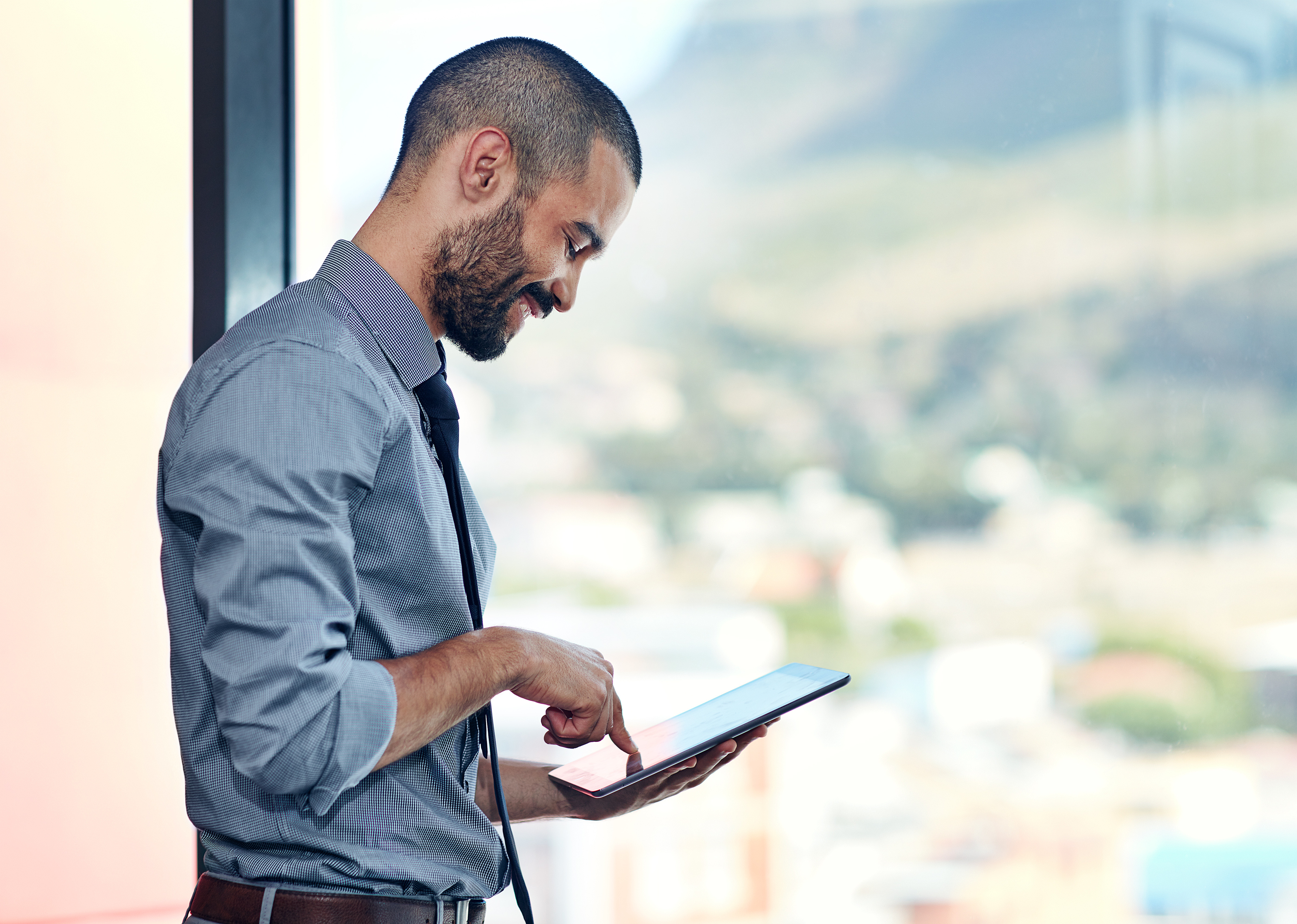 Smiling businessman in a dress shirt and tie using a tablet by a large window with a blurred city view.