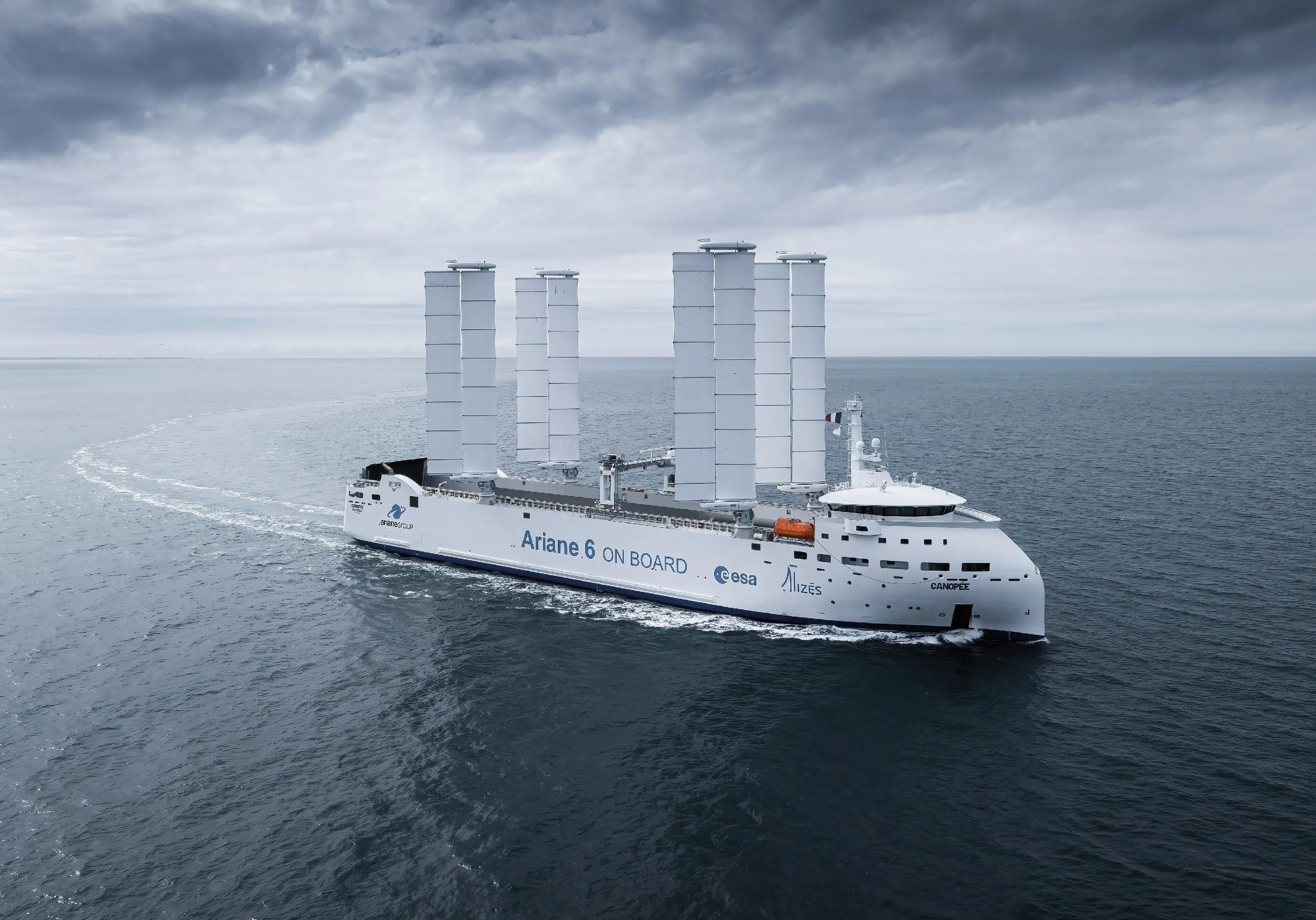 The Canopée Ro-Ro vessel equipped with four wing sails from OceanWings, on the ocean. The side of the vessel displays the text “Ariane 6 on board” along with logos of Alizés and ESA. 