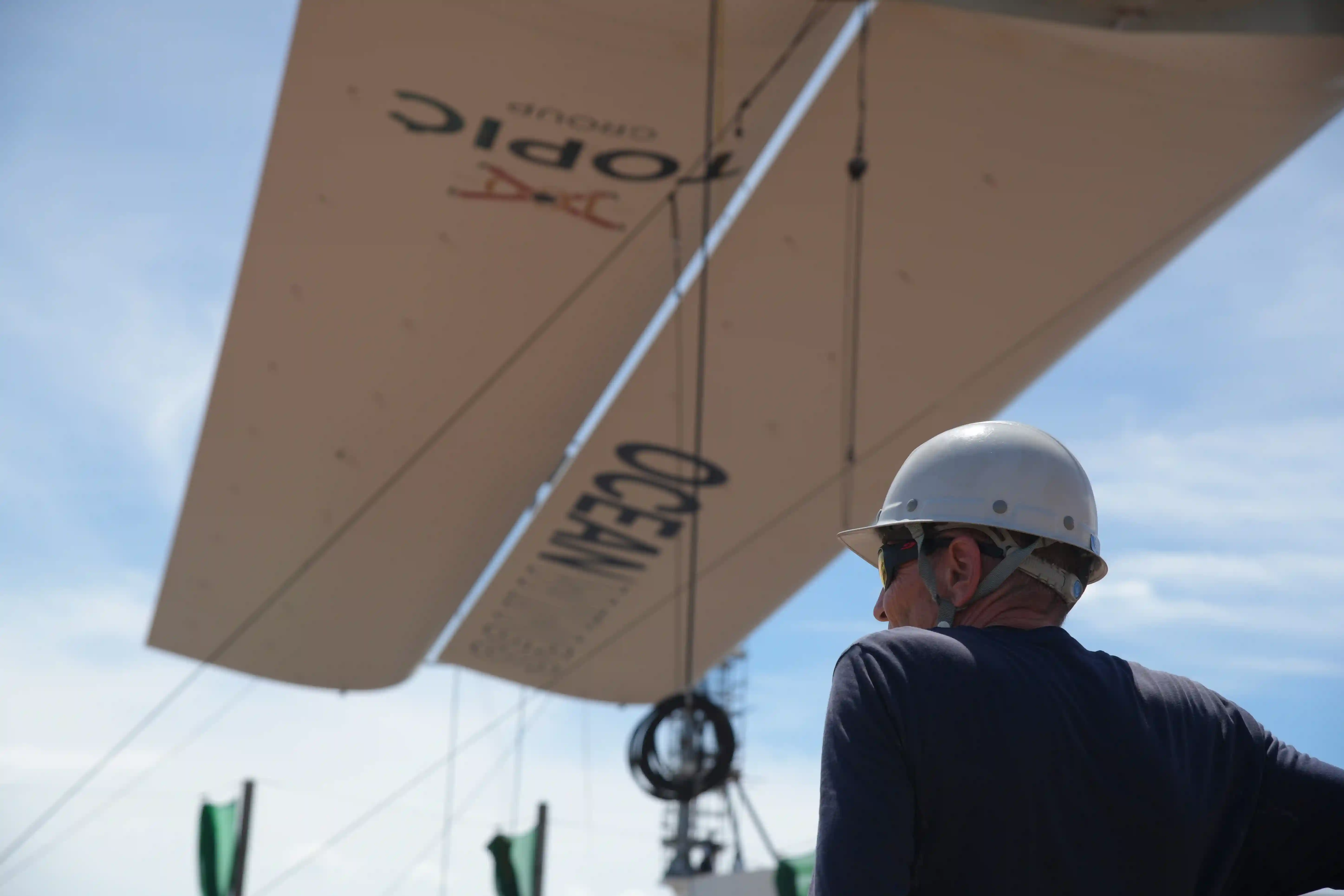 A white boom of a Lowerable Fixed Wingsail installed on a ship by several men at a port.