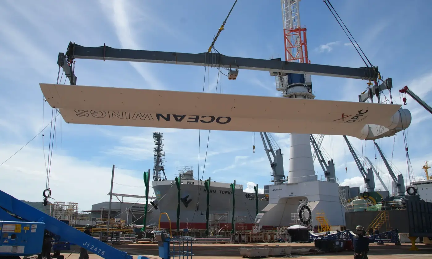 Several people are installing the boom of Lowerable Fixed Wingsail from OceanWings on the deck of the Ro-Ro Canopée vessel, at a port.
