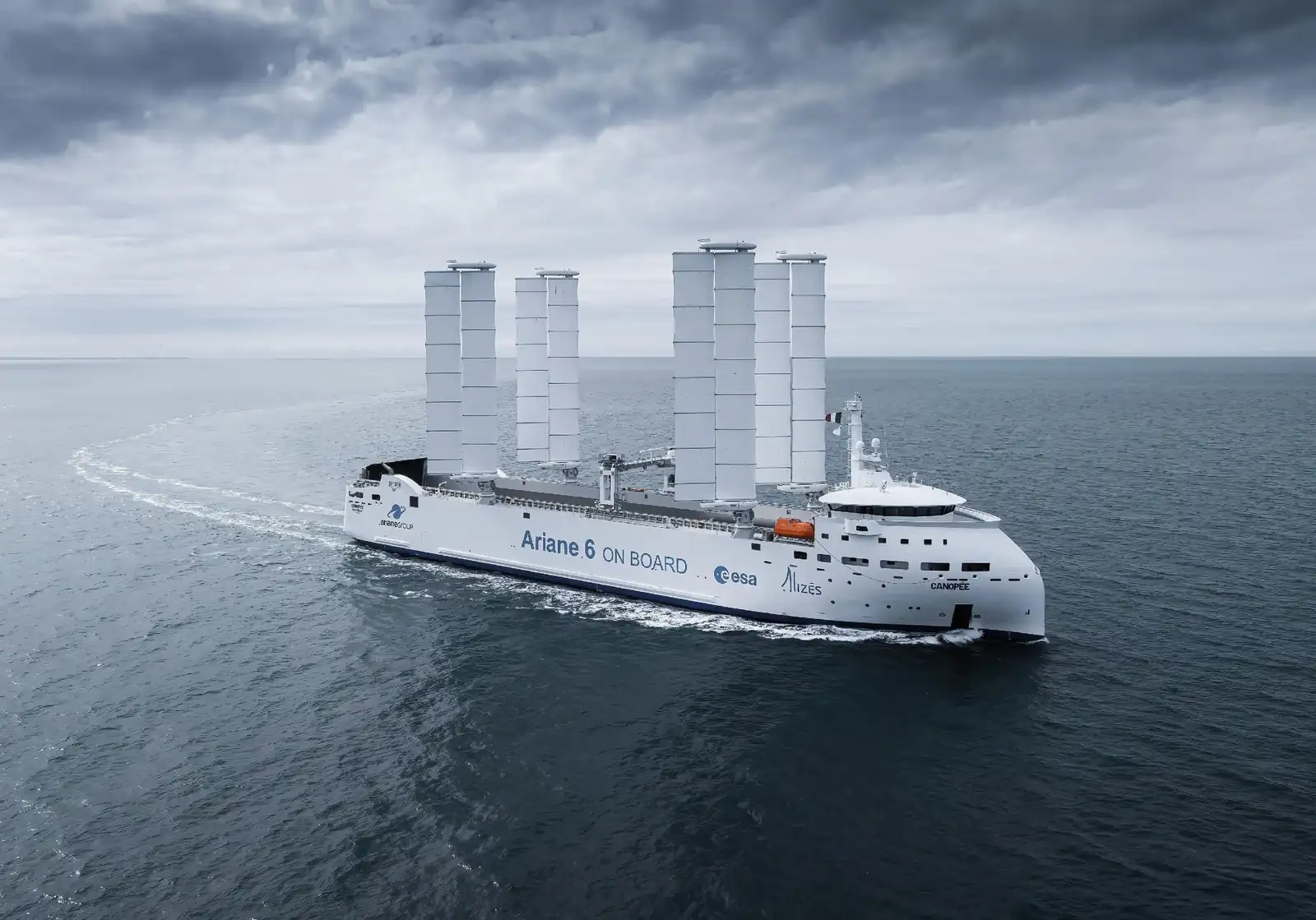 The Canopée Ro-Ro vessel equipped with four wing sails from OceanWings, on the ocean. The side of the vessel displays the text “Ariane 6 on board” along with logos of Alizés and ESA.