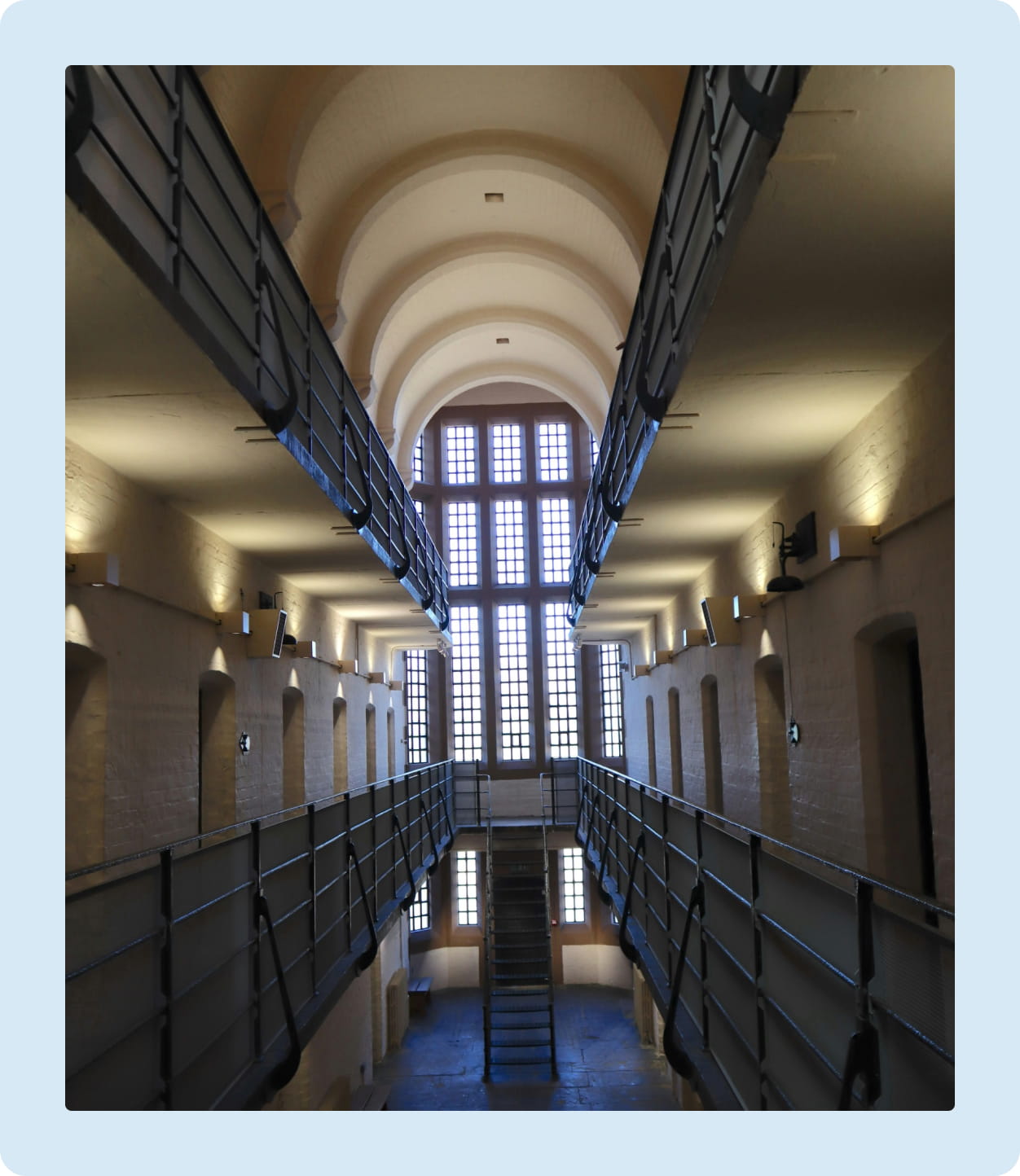 Interior view of a historic prison cell block with two levels of catwalks, arched ceiling, and large grid windows at the end.