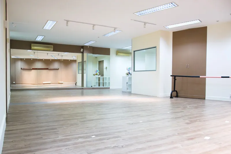 Empty dance studio with wooden floors, wall mirrors, ballet barre, and overhead fluorescent lights.
