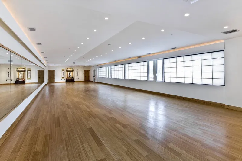 Spacious, empty room with wooden flooring, large wall mirror on the left, and illuminated rectangular windows on the right.