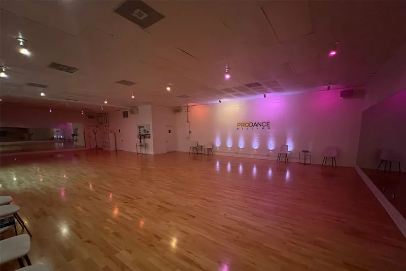 Spacious dance studio with wooden floor, wall mirrors, chairs, and colorful mood lighting.