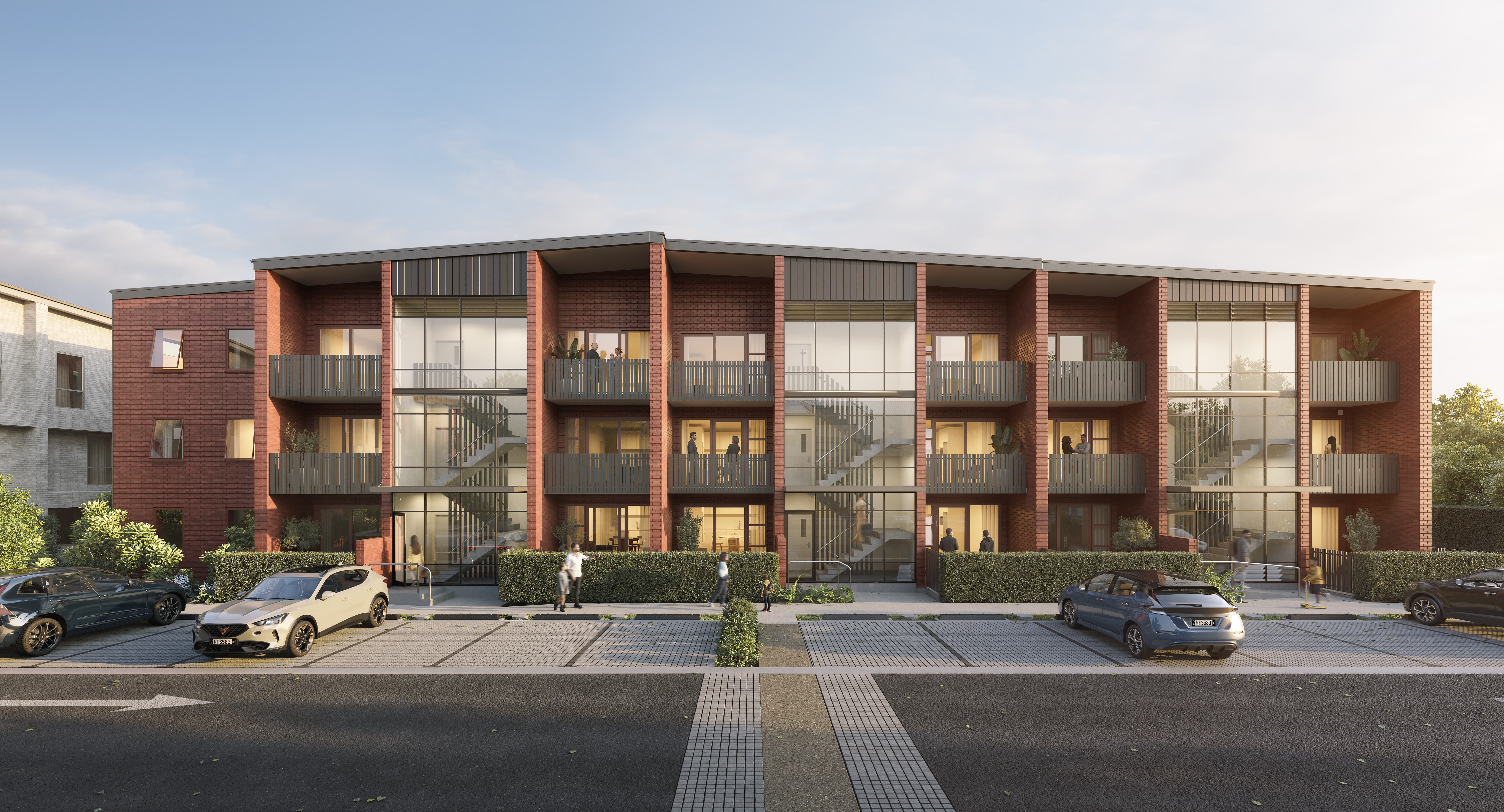 Modern three-story red brick apartment building with balconies, large glass stairwells, parked cars, and people walking on the sidewalk.