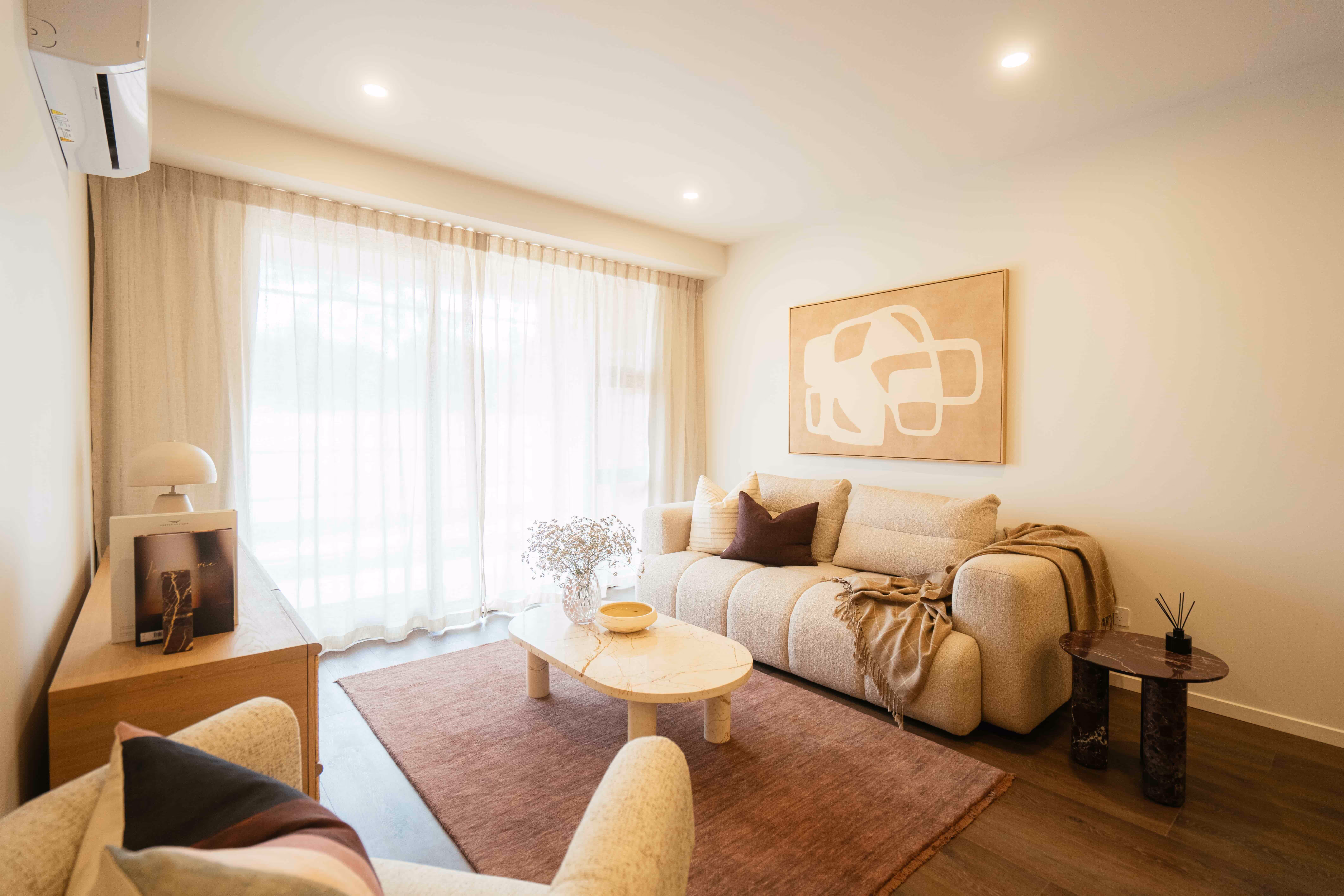 Cozy living room with a beige sofa, two pillows, a throw blanket, abstract wall art, a marble coffee table, a side table, and sheer curtains over large windows.