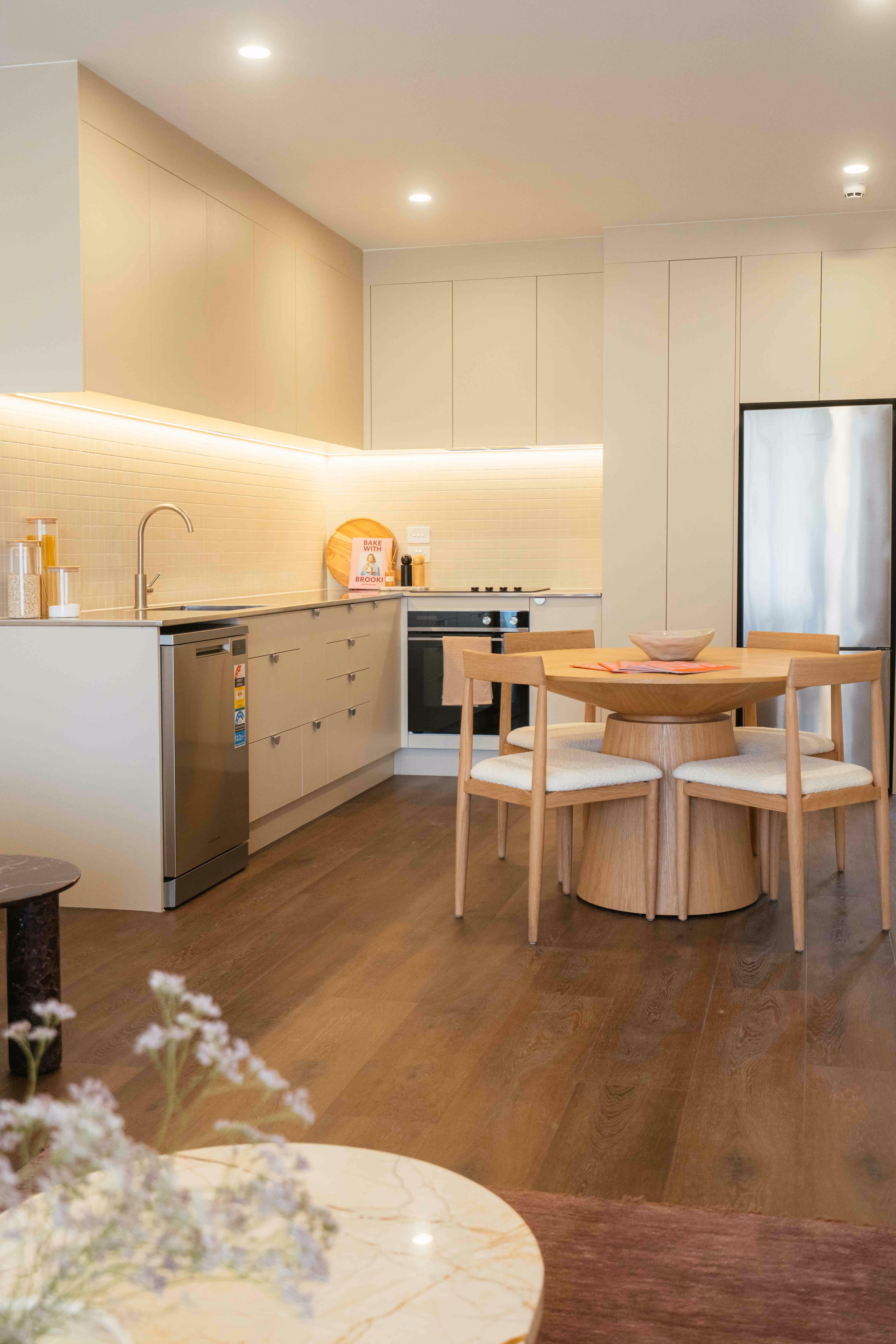 Modern kitchen with beige cabinetry, under-cabinet lighting, stainless steel appliances, and a round wooden dining table with four chairs.