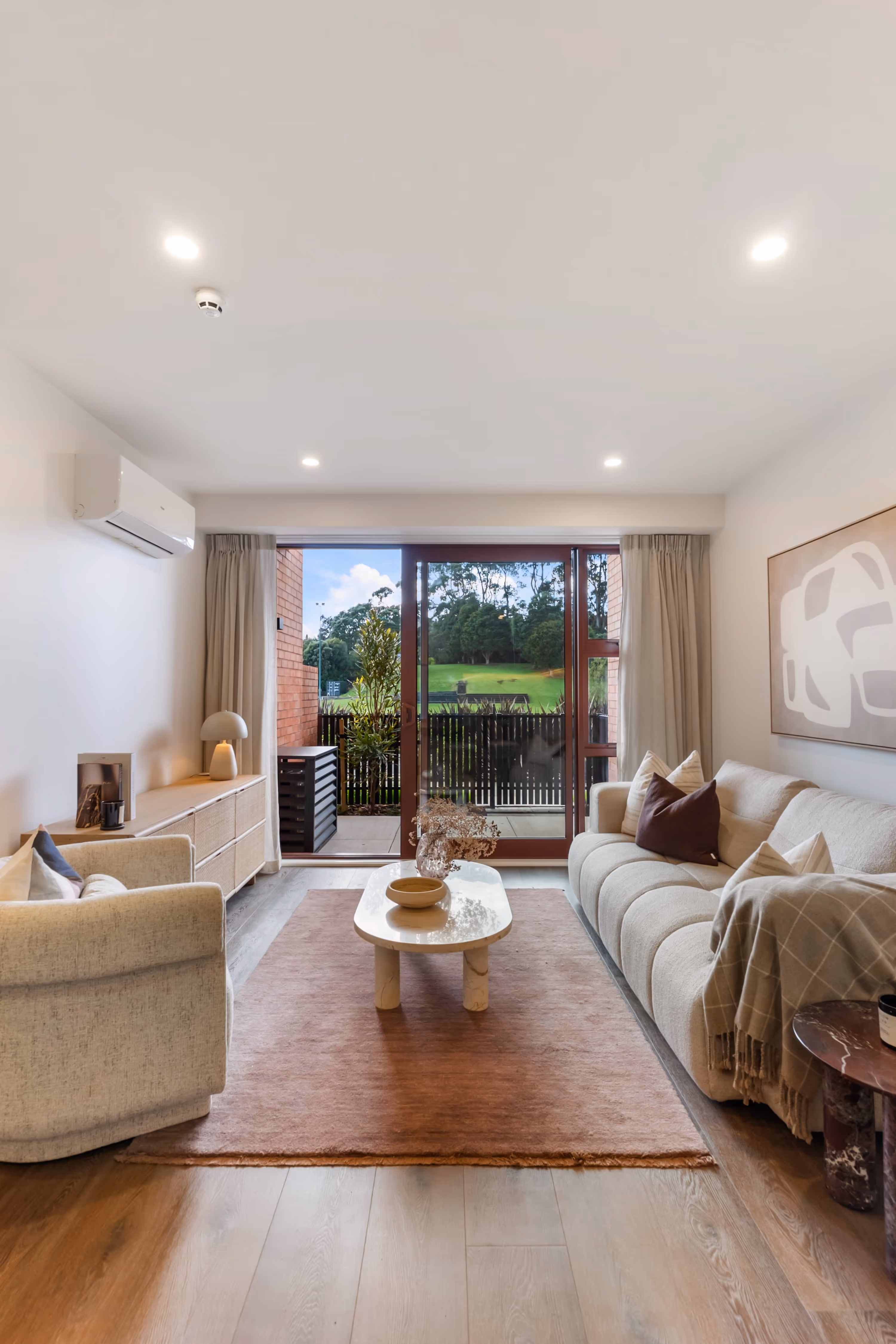 Living room with beige sofa, armchair, coffee table on rug, and sliding glass doors opening to green outside.