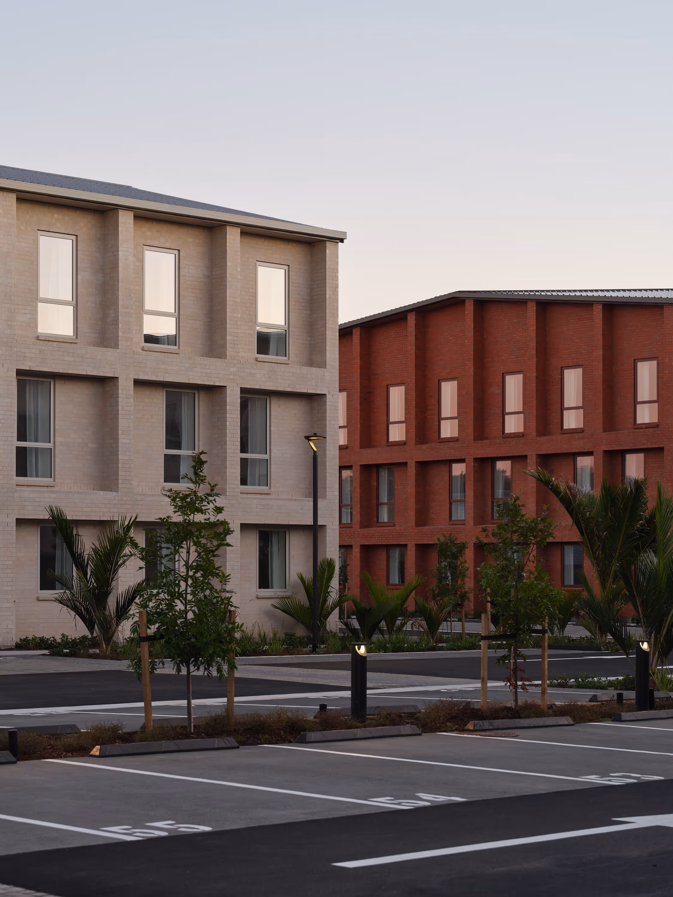 Empty parking lot with marked spaces in front of modern beige and red brick buildings with trees.