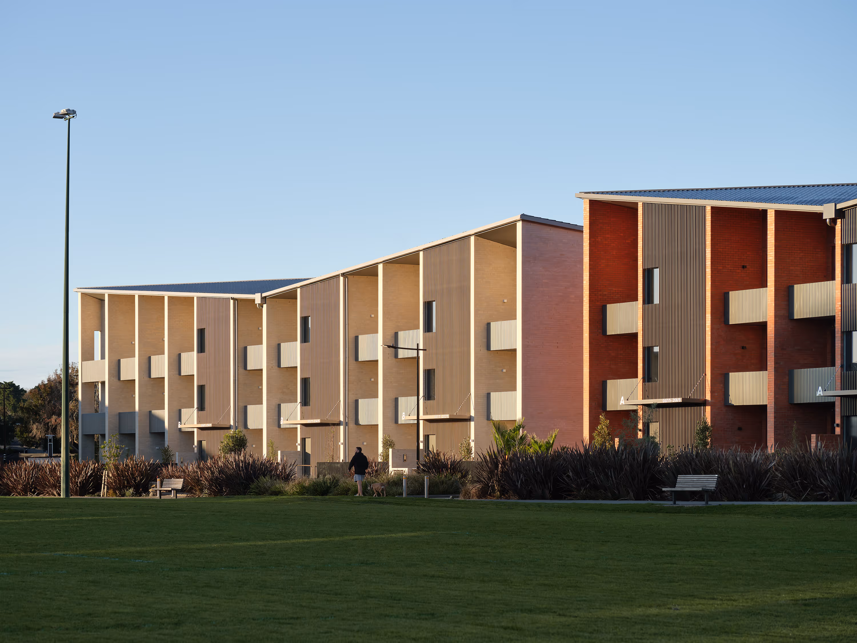 Modern apartment buildings with beige and red walls, balconies, and green lawn in front.