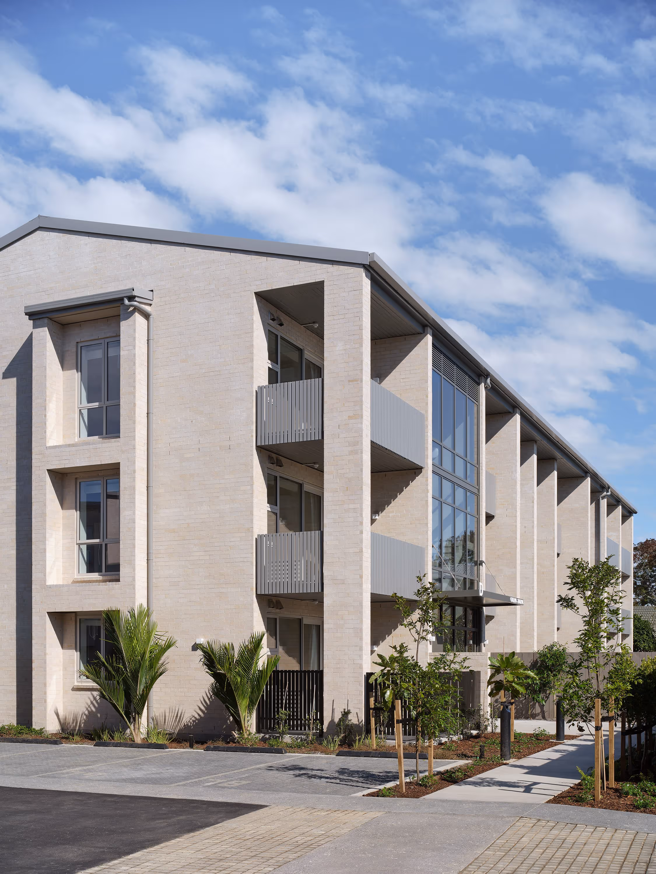 Modern three-story apartment building with balconies, large windows, plants, and a clear blue sky.