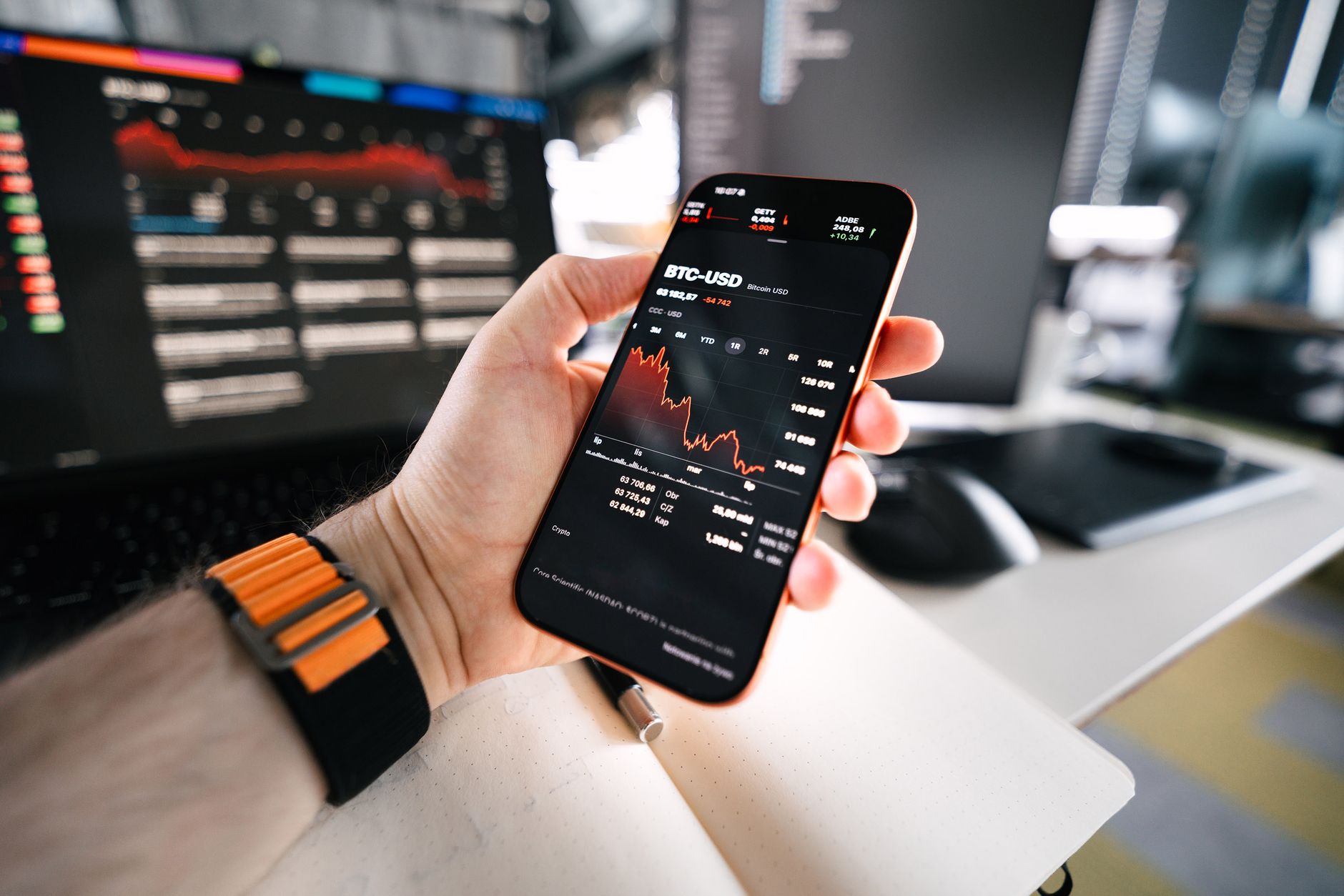 Mano sosteniendo un smartphone con el gráfico de precio de Bitcoin, con una computadora de fondo