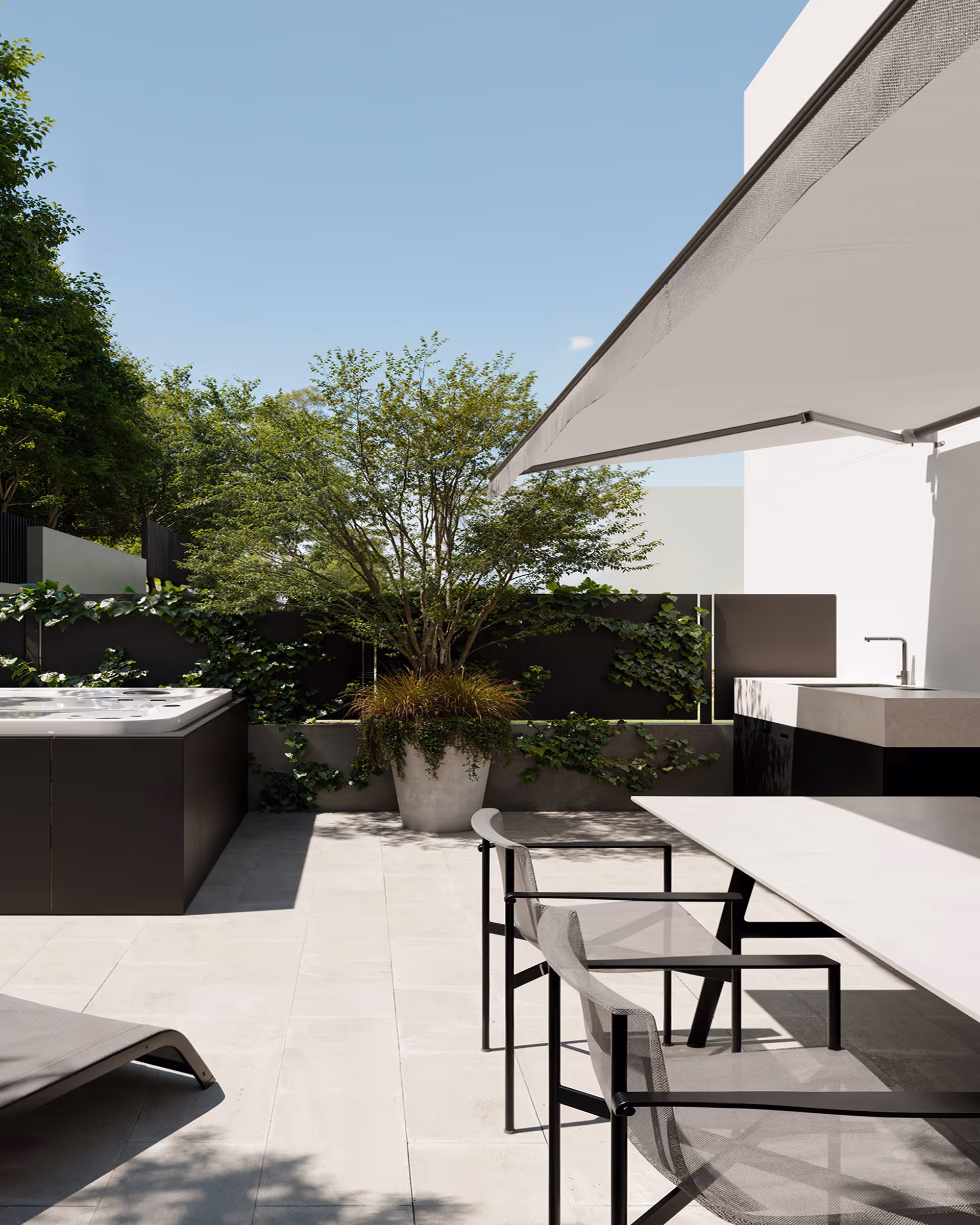 Modern outdoor patio with sunshade, two mesh chairs, a white table, a black hot tub, and a large potted tree against a black fence.