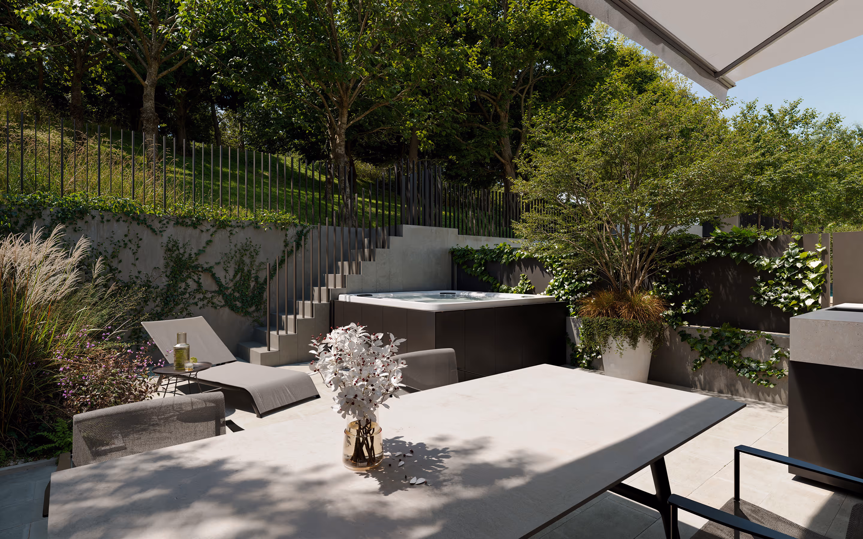 Modern outdoor patio with a beige table, mesh chairs, a chaise lounge, potted plants, and a hot tub surrounded by greenery.