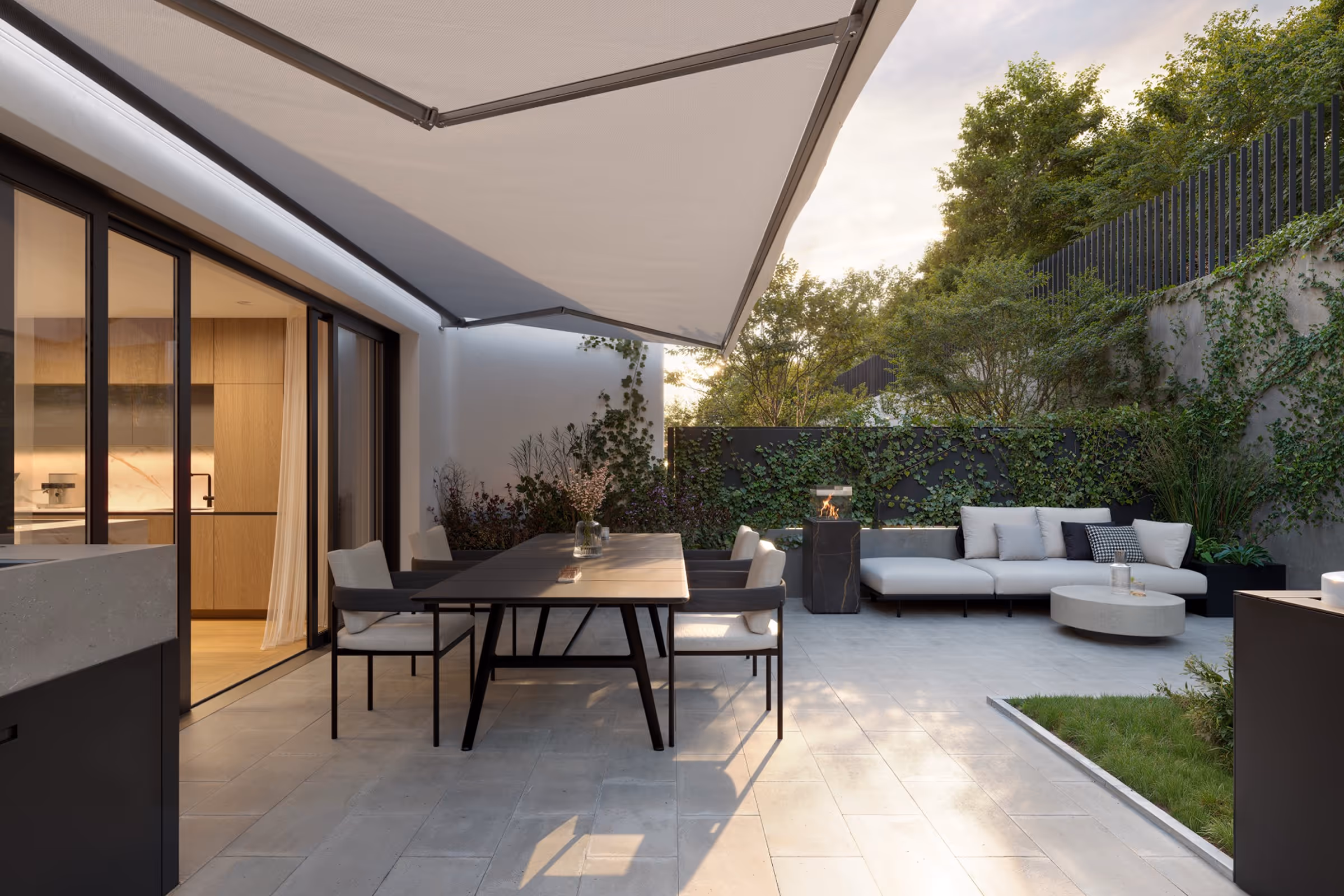 Modern outdoor patio with dining table, chairs, white sofa, coffee table, and greenery under an awning.