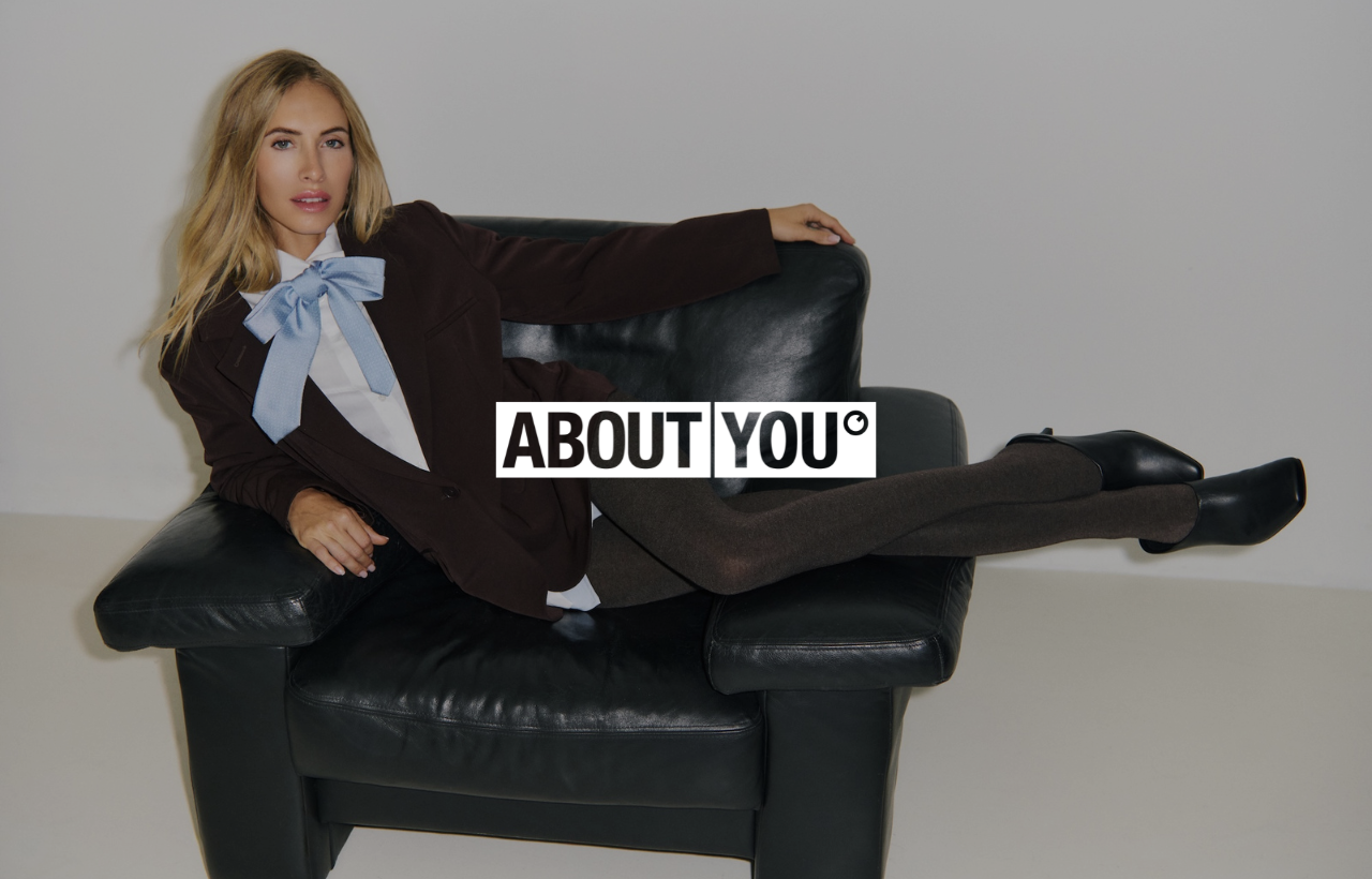 Person sitting on a black sofa holding a large ‘ABOUT YOU’ sign, used to illustrate the new Sello integration with the About You marketplace.