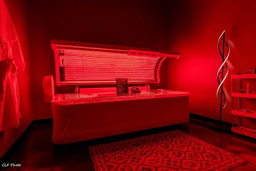 Red-lit room with an open tanning bed, a decorative rug on the floor, a modern floor lamp, and shelves in the corner.